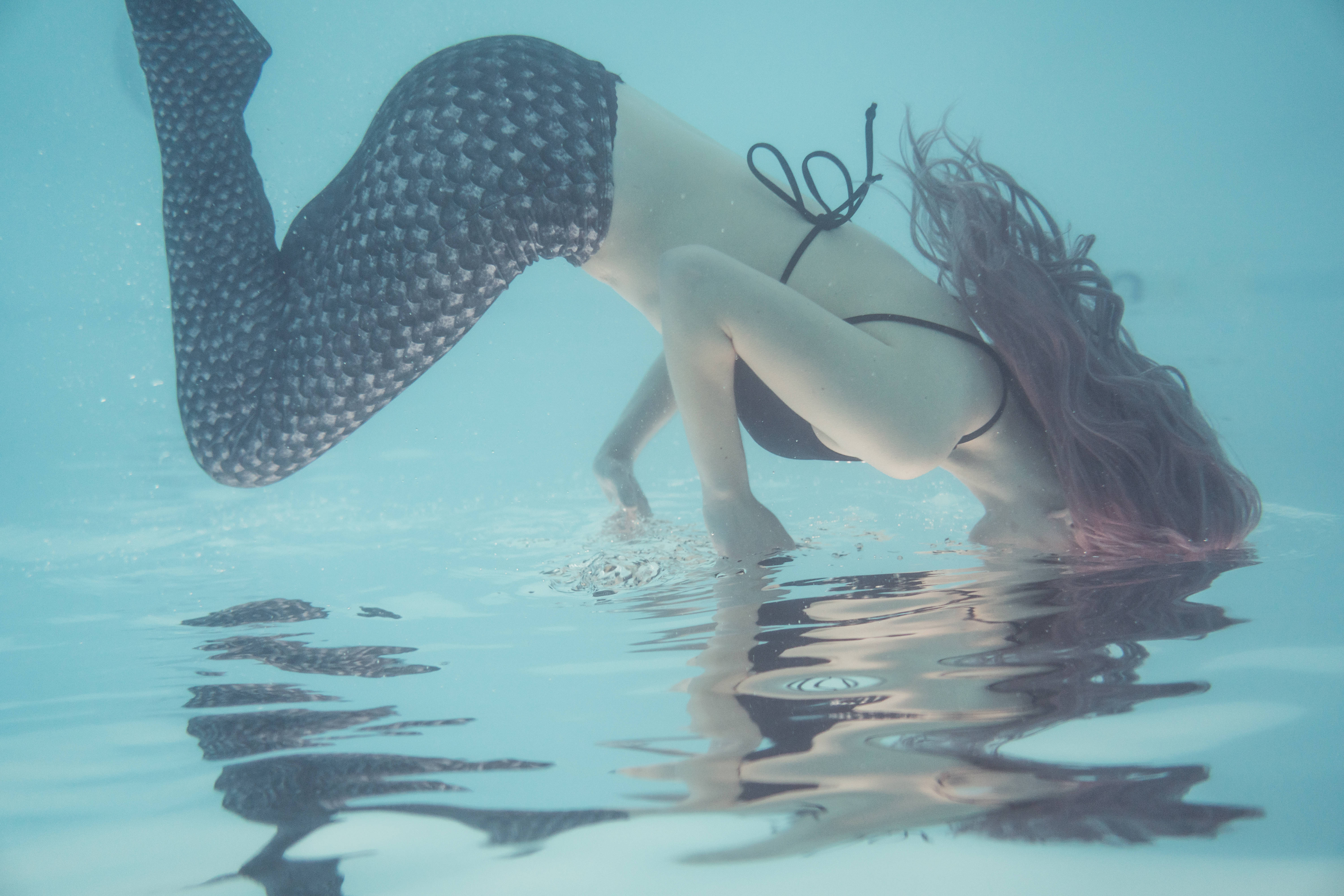 The Little Mermaid 桌布 壁紙 — 一位穿著黑色鱗片圖案美人魚尾巴的女性，頭髮飄逸，優雅地潛入水中，在漣漪的水面上形成寧靜的倒影。