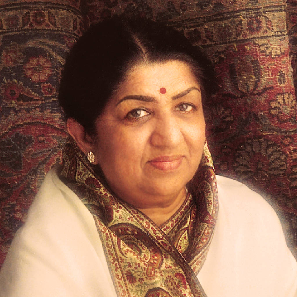 Lata Mangeshkar 桌布 壁紙 — 一位端莊的女性，帶有紅色眉心貼和華麗的圍巾，在豐富圖案的掛毯背景下柔和地凝視。