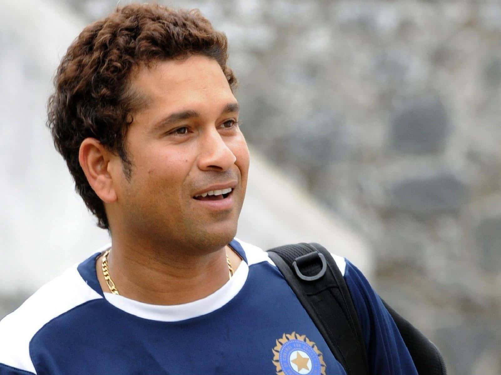 Sachin Tendulkar 桌布 壁紙 — 一位笑容滿面、棕色捲髮的男子，身穿藍色印度板球球衣，背著黑色背包，背景是模糊的石牆。