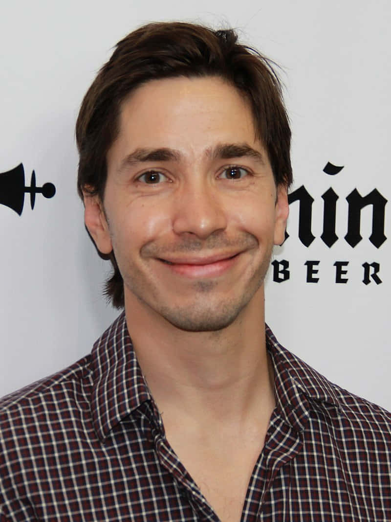 Justin Long 桌布 壁紙 — 一位笑容滿面、棕色頭髮、穿著格紋襯衫的男士站在白色背景前，背景有黑色文字和標誌。