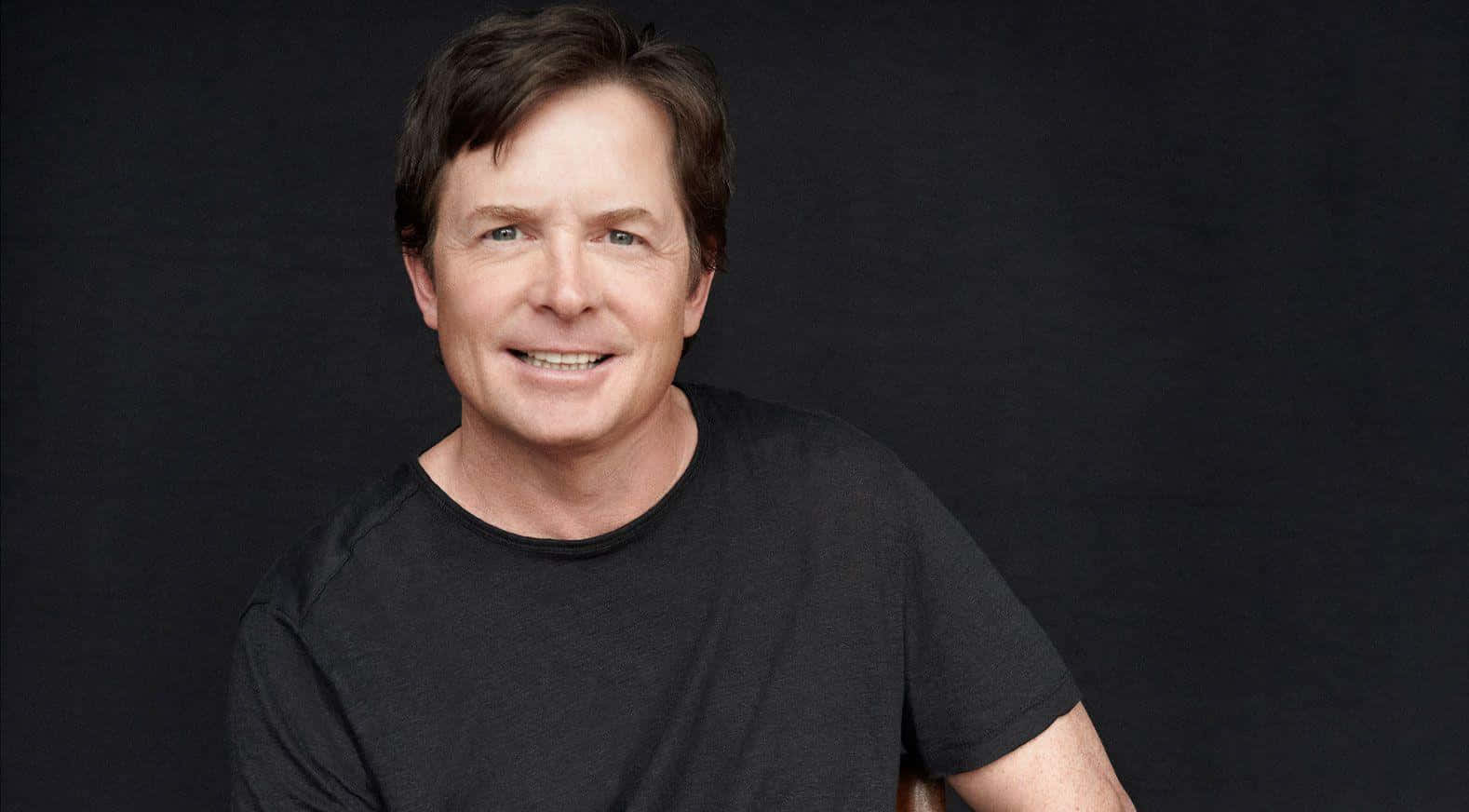 michael j fox 桌布 壁紙 — 一位笑容滿面、棕色頭髮的男子，在深色攝影棚背景前穿著黑色T恤。