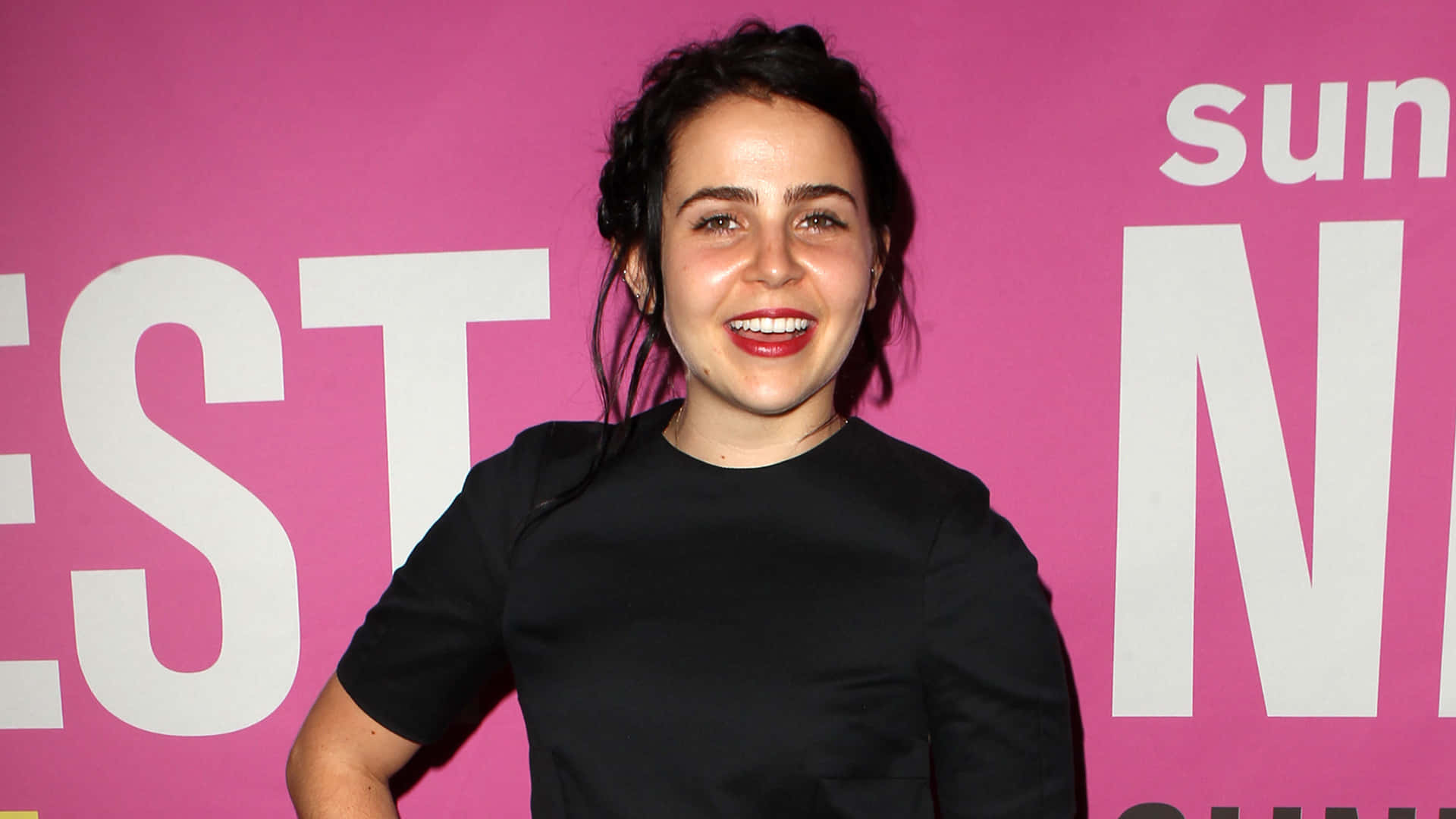 Mae Whitman 桌布 壁紙 — 一位笑容滿面、深色頭髮、塗著紅色唇膏的女性自信地站在明亮的粉紅色背景前，背景上有白色文字。
