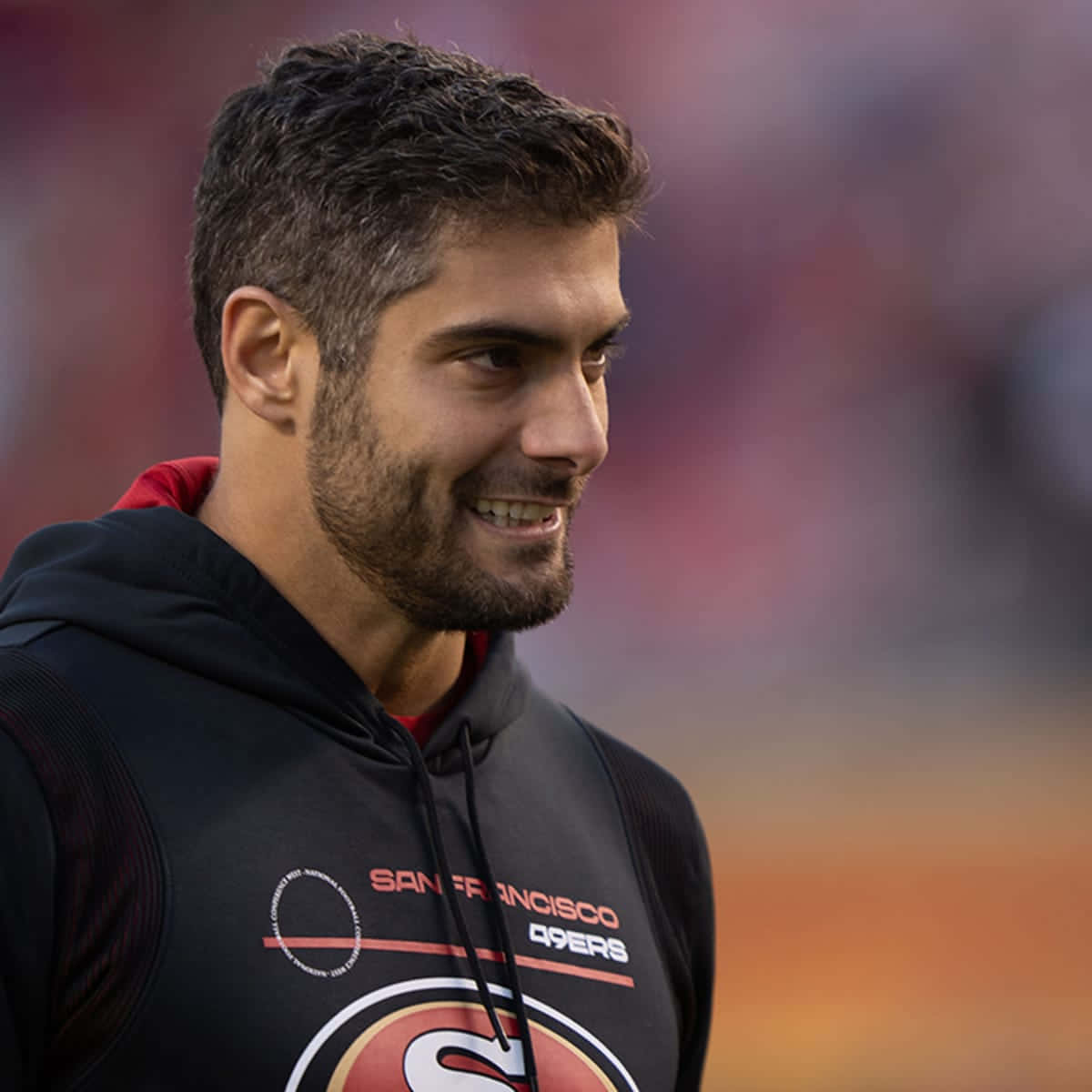 Jimmy Garoppolo 桌布 壁紙 — 一位笑容滿面、深色頭髮和鬍鬚的男子，在模糊的體育場背景下穿著黑色舊金山49人連帽衫。