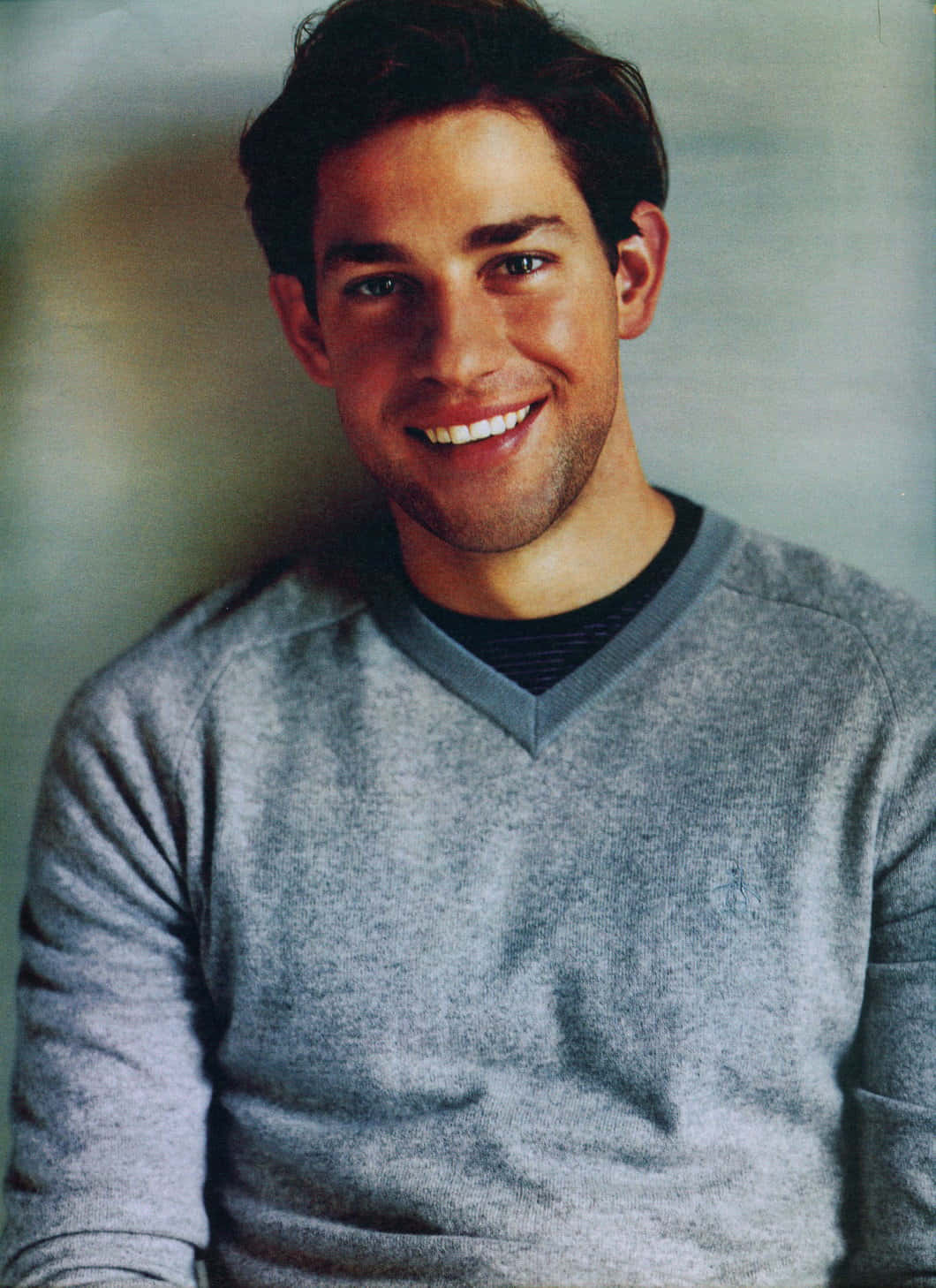 john krasinski 桌布 壁紙 — 一位笑容滿面、深色頭髮的男子，穿著灰色V領毛衣，背景柔和模糊。
