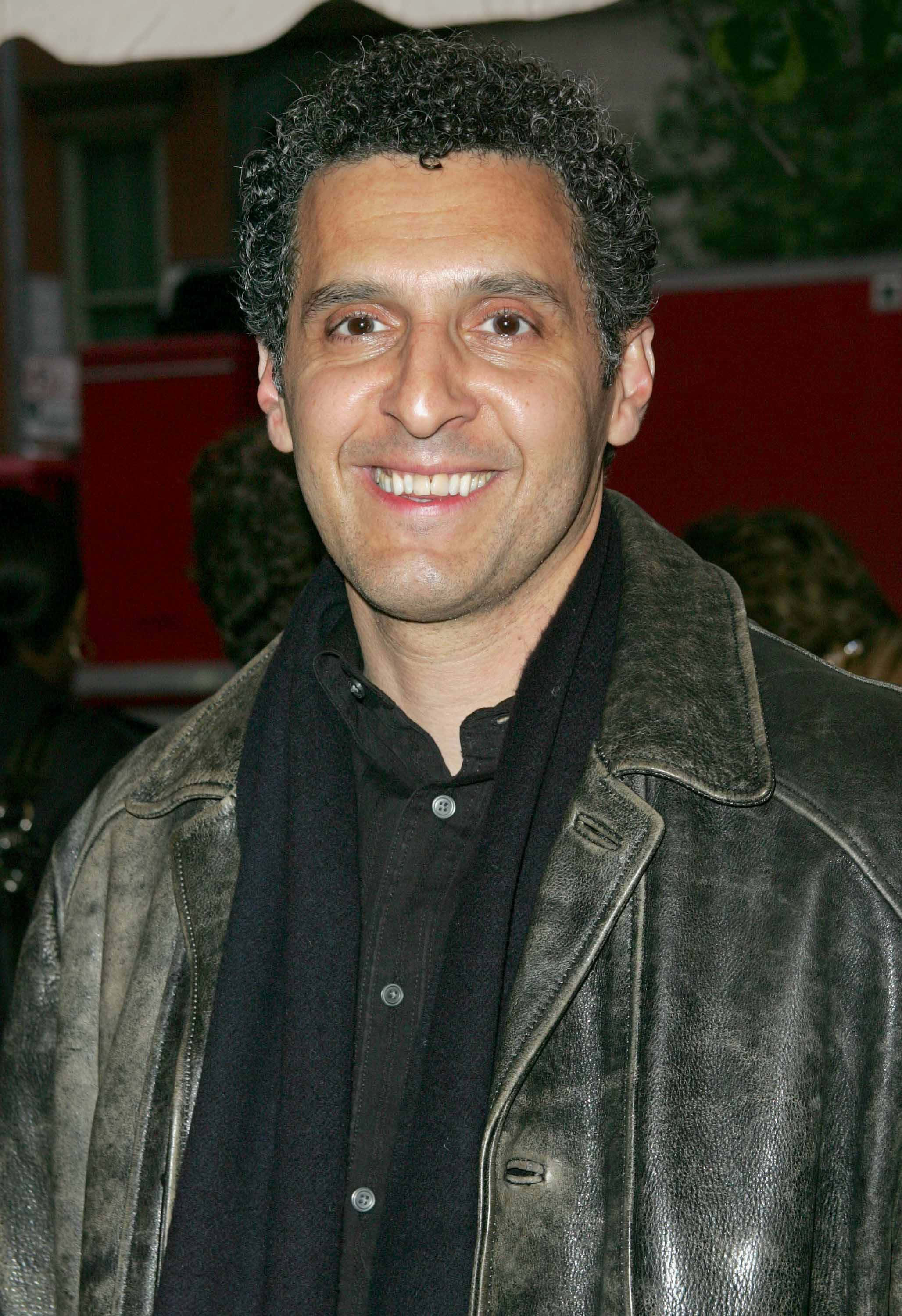 John Turturro 桌布 壁紙 — 一位笑容滿面、留著深色捲髮的男子，身穿一件破舊的棕色皮夾克，內搭黑色襯衫和圍巾。