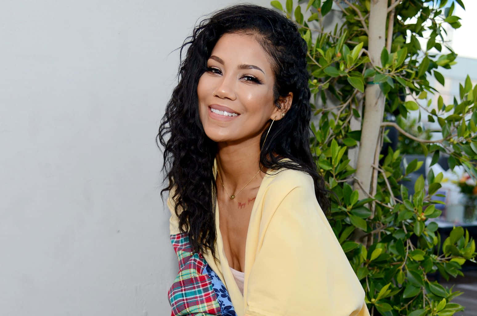 jhene aiko 桌布 壁紙 — 一位笑容滿面、留著深色長捲髮的女性，身穿黃色開襟毛衣和格紋上衣，背景是白色牆壁和綠色植物。