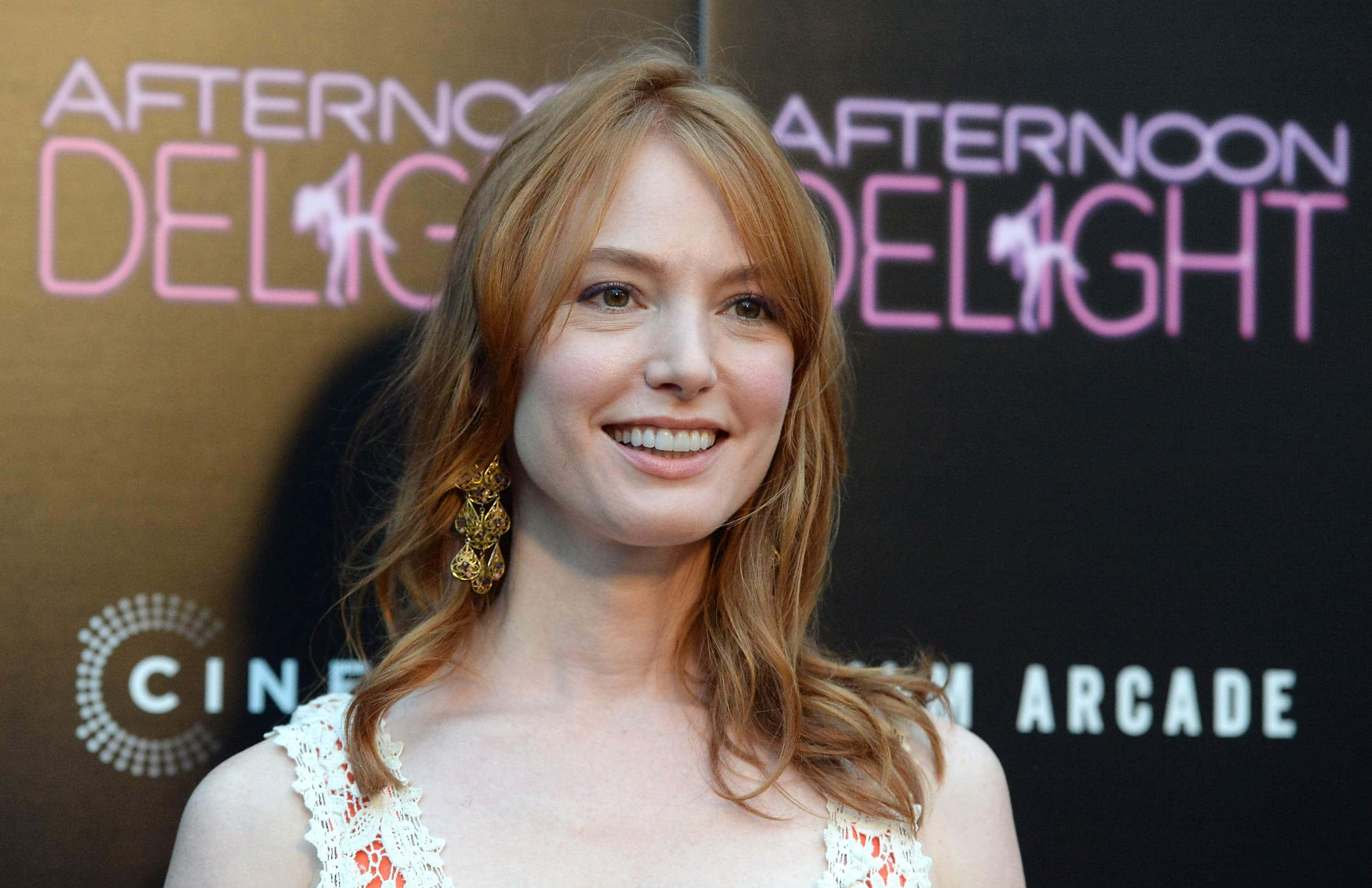 Alicia Witt 桌布 壁紙 — 一位笑容滿面、紅髮金耳環的女性在粉紅色「Afternoon Delight」活動背景前擺姿勢。