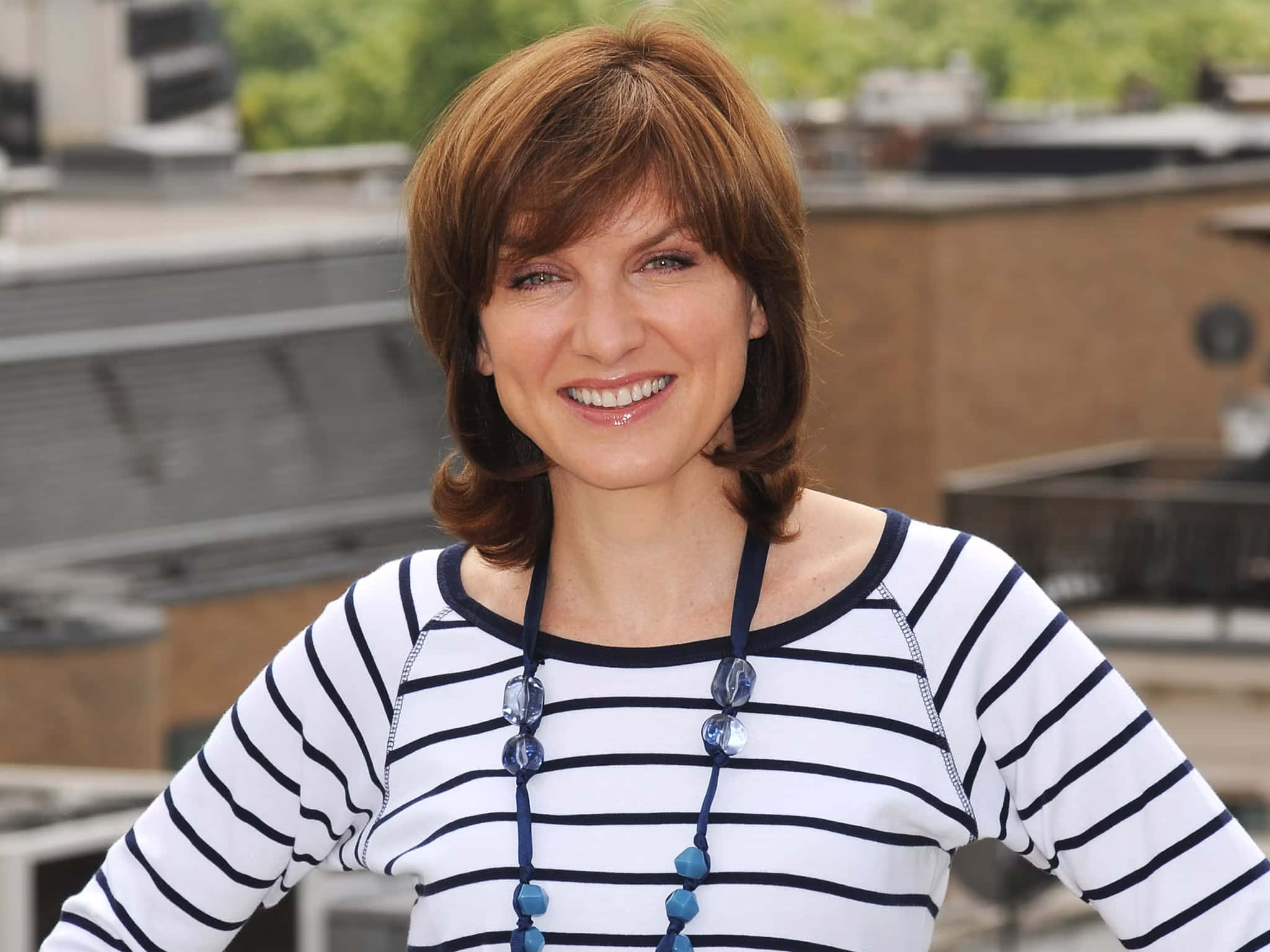 Fiona Bruce 桌布 壁紙 — 一位笑容滿面的棕髮女性，身穿海軍藍與白色條紋上衣，背景是模糊的城市屋頂。