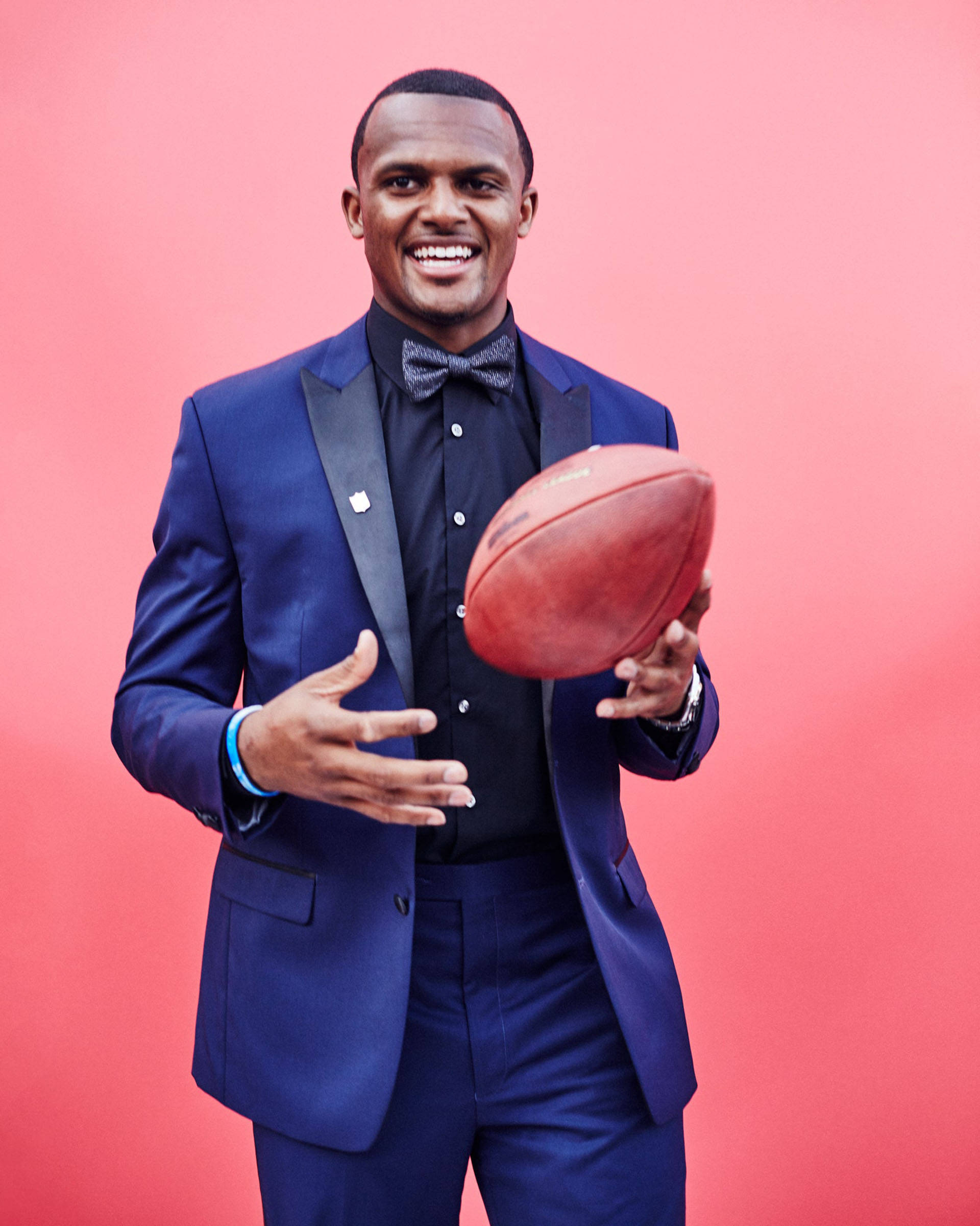 Deshaun Watson 桌布 壁紙 — 一位笑容滿面的男士身穿筆挺藍色西裝，在充滿活力的粉紅色攝影棚背景前抱著一個足球。