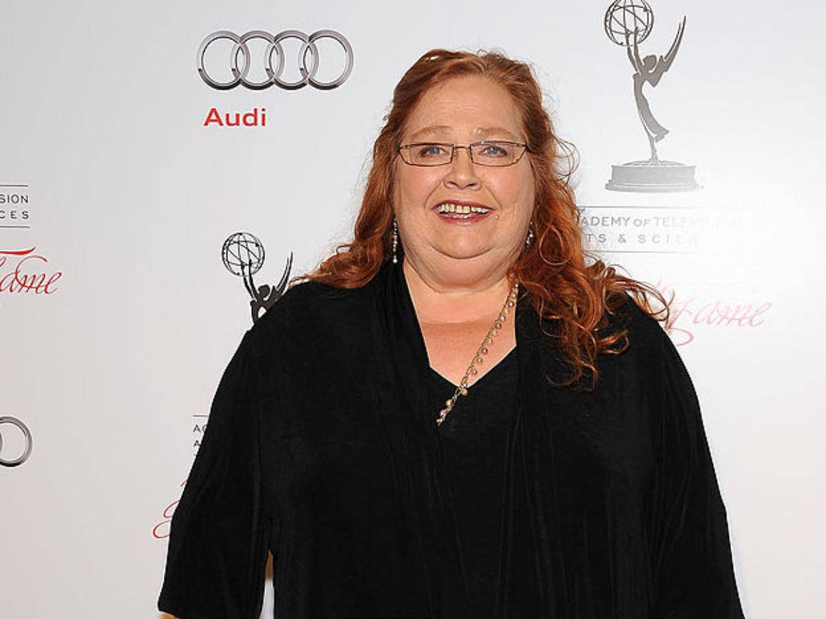 女演員 桌布 壁紙 — 一位笑容滿面的紅髮戴眼鏡女士，在背後有Audi和Emmy標誌的頒獎活動中穿著黑色天鵝絨上衣。
