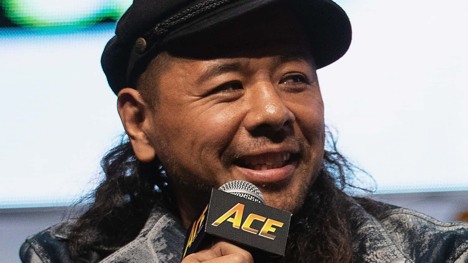 interview 桌布 壁紙 — 一位笑容滿面的長黑髮男子戴著黑色帽子，在活動中手持印有「ACE」標誌的麥克風。