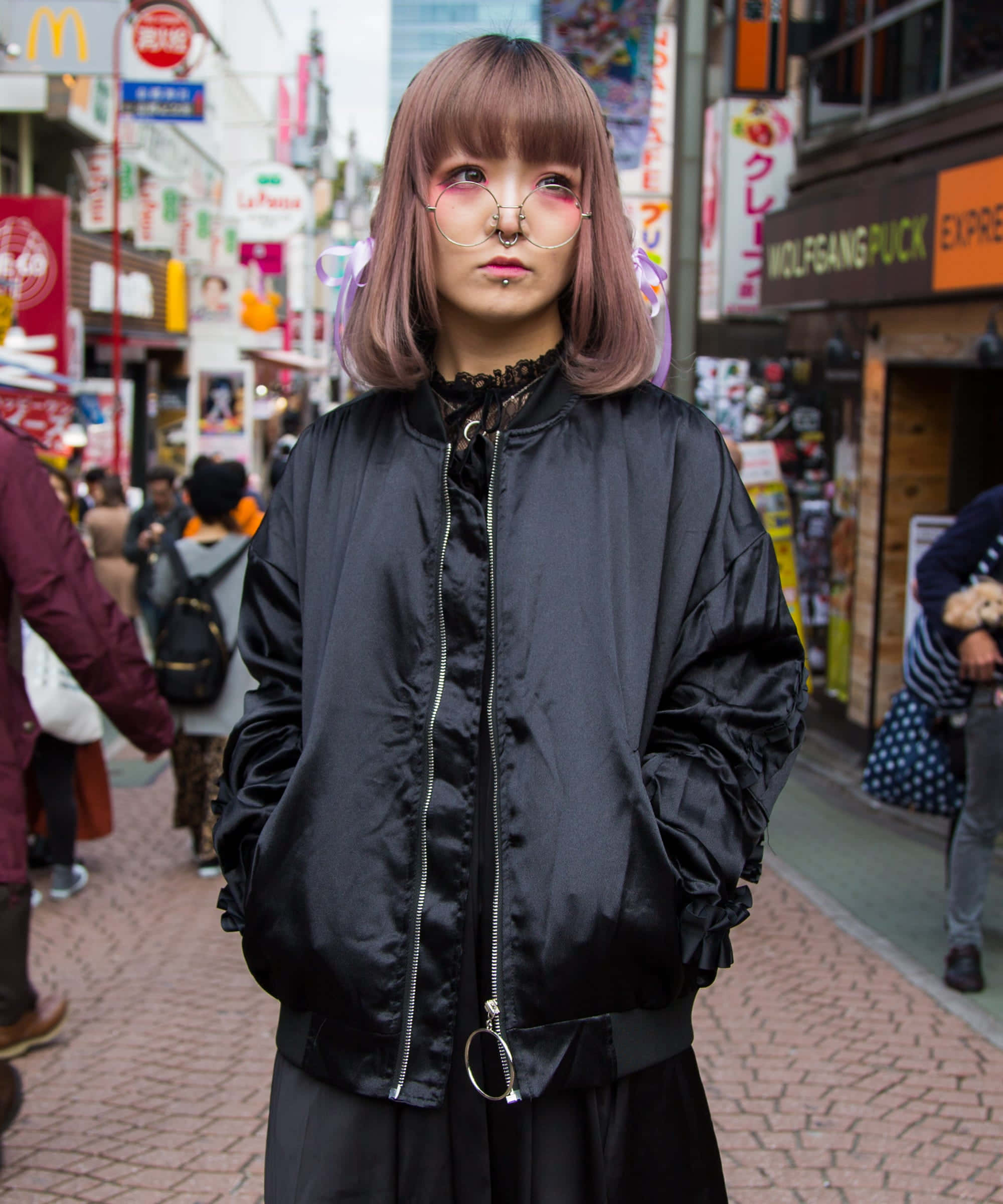 Harajuku Fashion 桌布 壁紙 — 一位粉紅色頭髮、戴眼鏡的年輕女性自信地站在東京一條繁華的街道上，街道兩旁是色彩繽紛的招牌和商店。