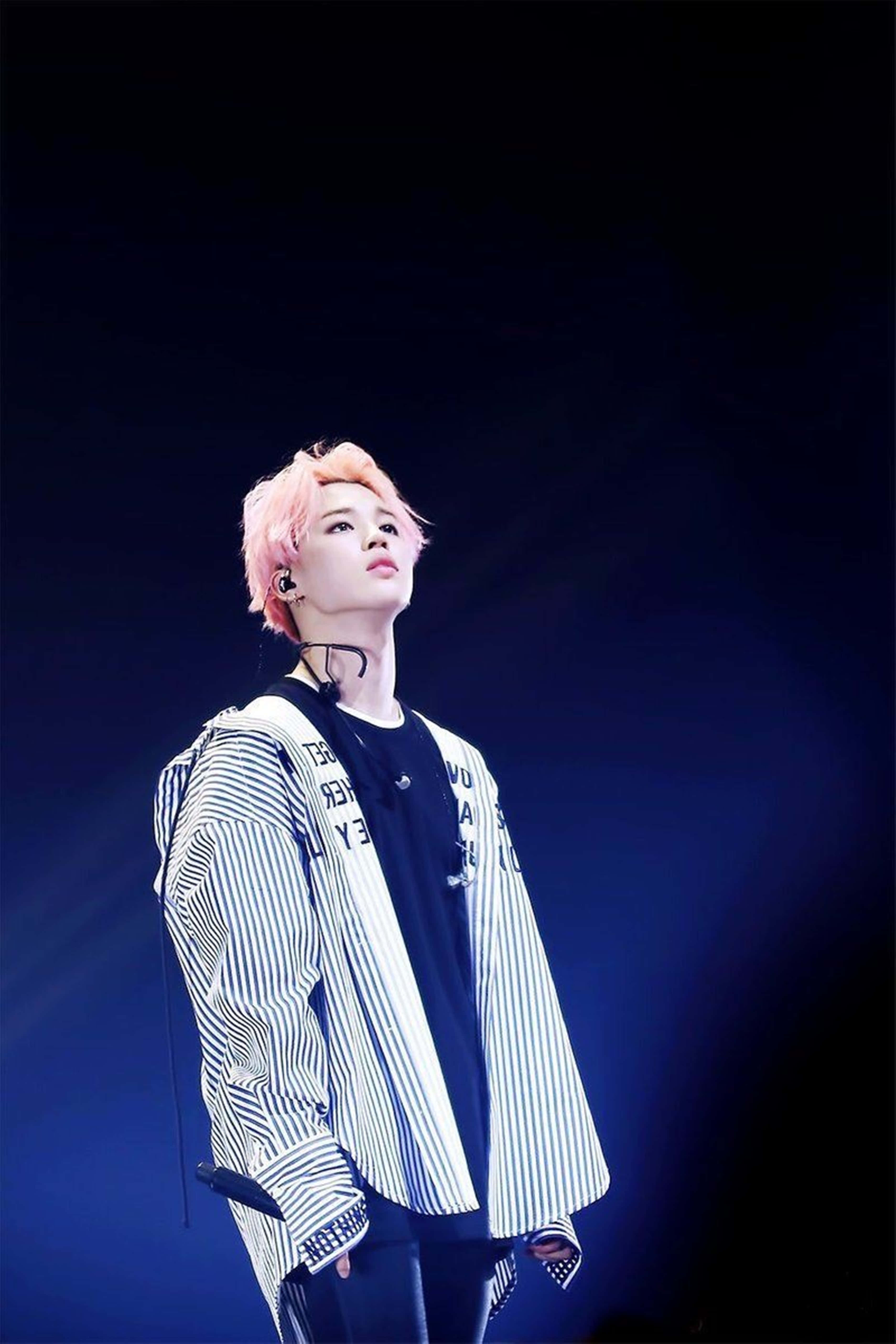 Jimin Bts Cute 桌布 壁紙 — 一位粉紅色頭髮、身穿條紋外套的表演者，在黑暗背景下被藍色舞台燈光照亮。