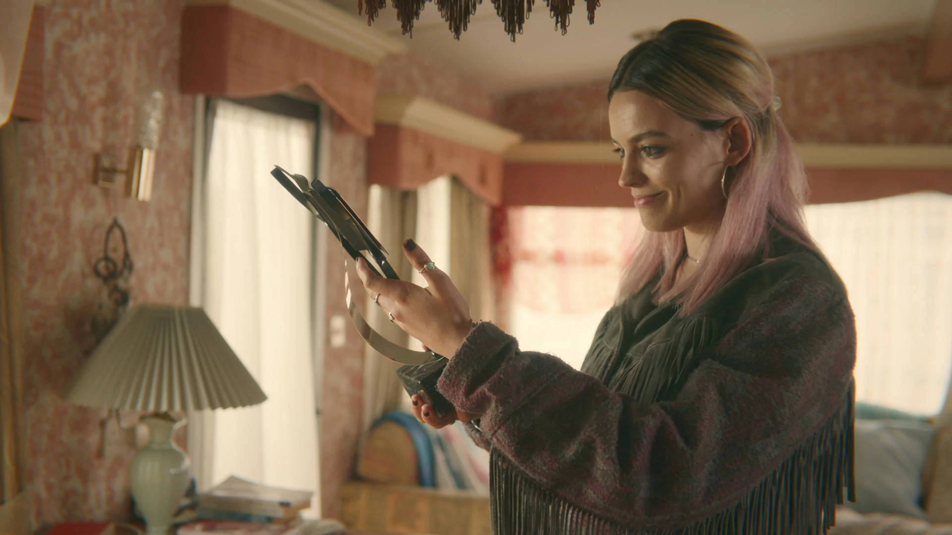 maeve wiley 桌布 壁紙 — 一位粉紅色頭髮的女性在溫暖燈光的復古房間裡微笑著拿著平板電腦，房間裡有花卉壁紙。