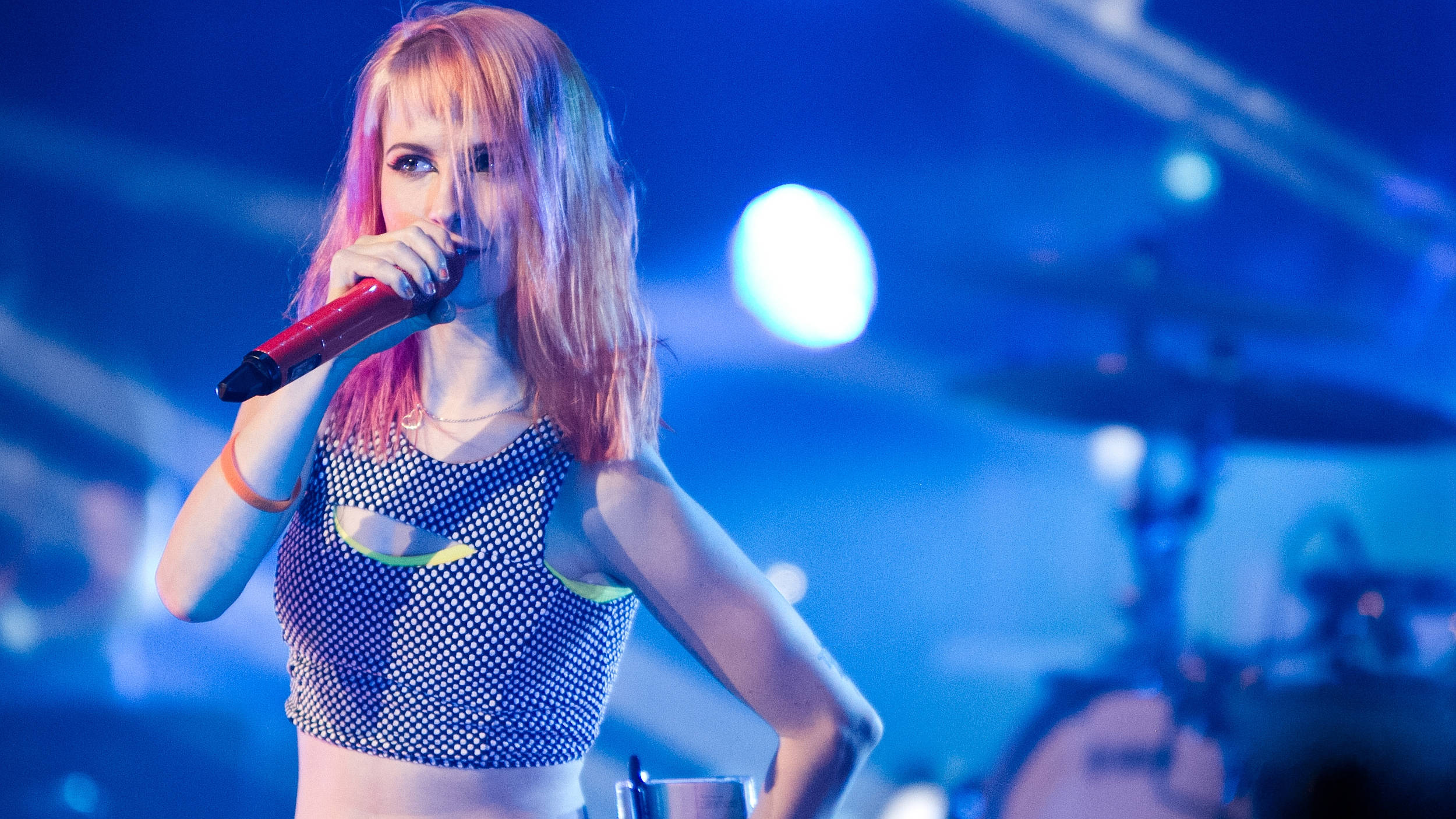 Hayley Williams 桌布 壁紙 — 一位粉紅色頭髮的女歌手在藍色舞台燈光下熱情地對著紅色麥克風唱歌，背景有鼓組。
