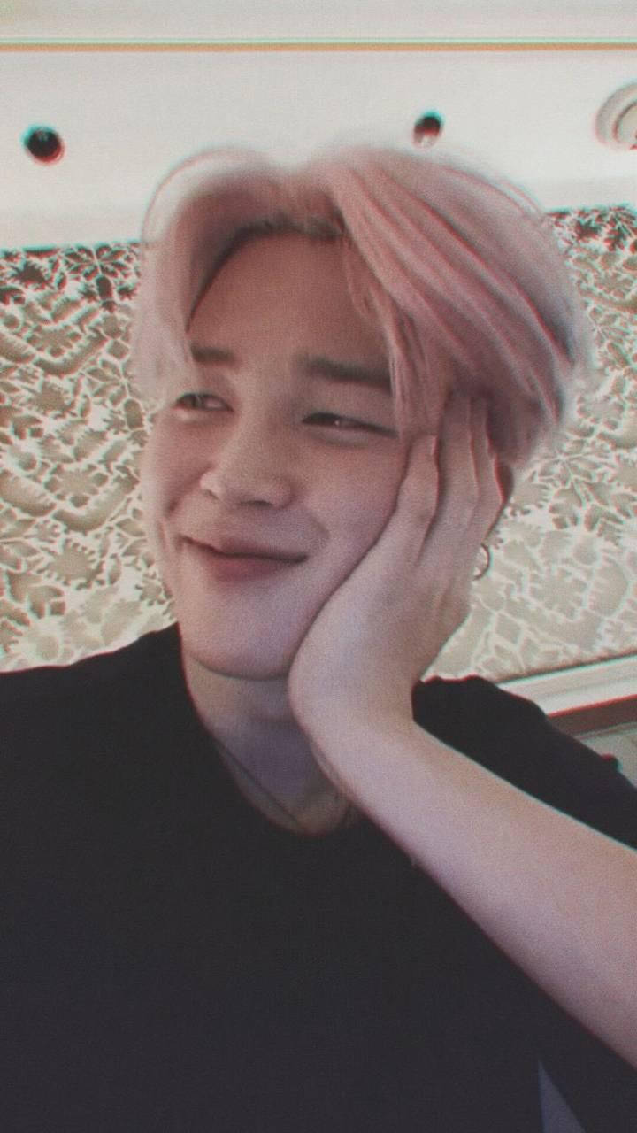 Jimin Aesthetic 桌布 壁紙 — 一位粉紅色頭髮的年輕男子微笑著，將臉頰靠在手上，背景是圖案壁紙。