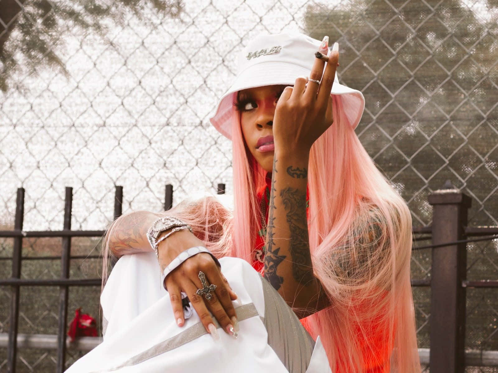 Rico Nasty 桌布 壁紙 — 一位粉紅頭髮和刺青的人自信地在鐵絲網前擺姿勢，戴著白色漁夫帽和分層首飾。