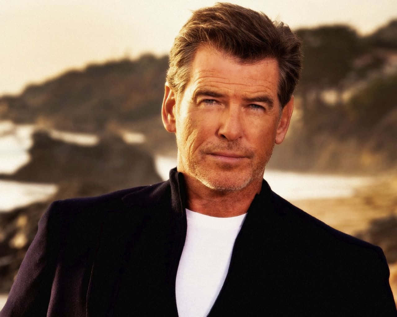 pierce brosnan 桌布 壁紙 — 一位粗獷英俊、頭髮花白的男士自信地站在陽光普照的海岸懸崖背景下。