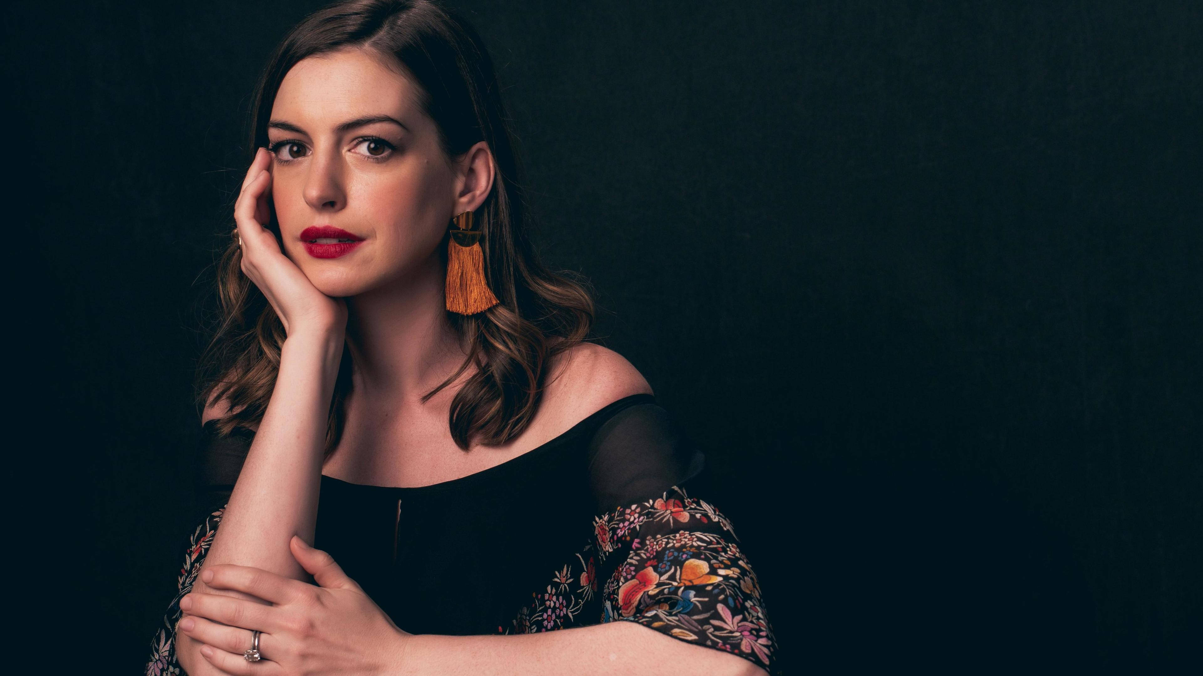 Anne Hathaway 桌布 壁紙 — 一位紅唇、戴流蘇耳環的女性，在深色背景下將下巴擱在手上。