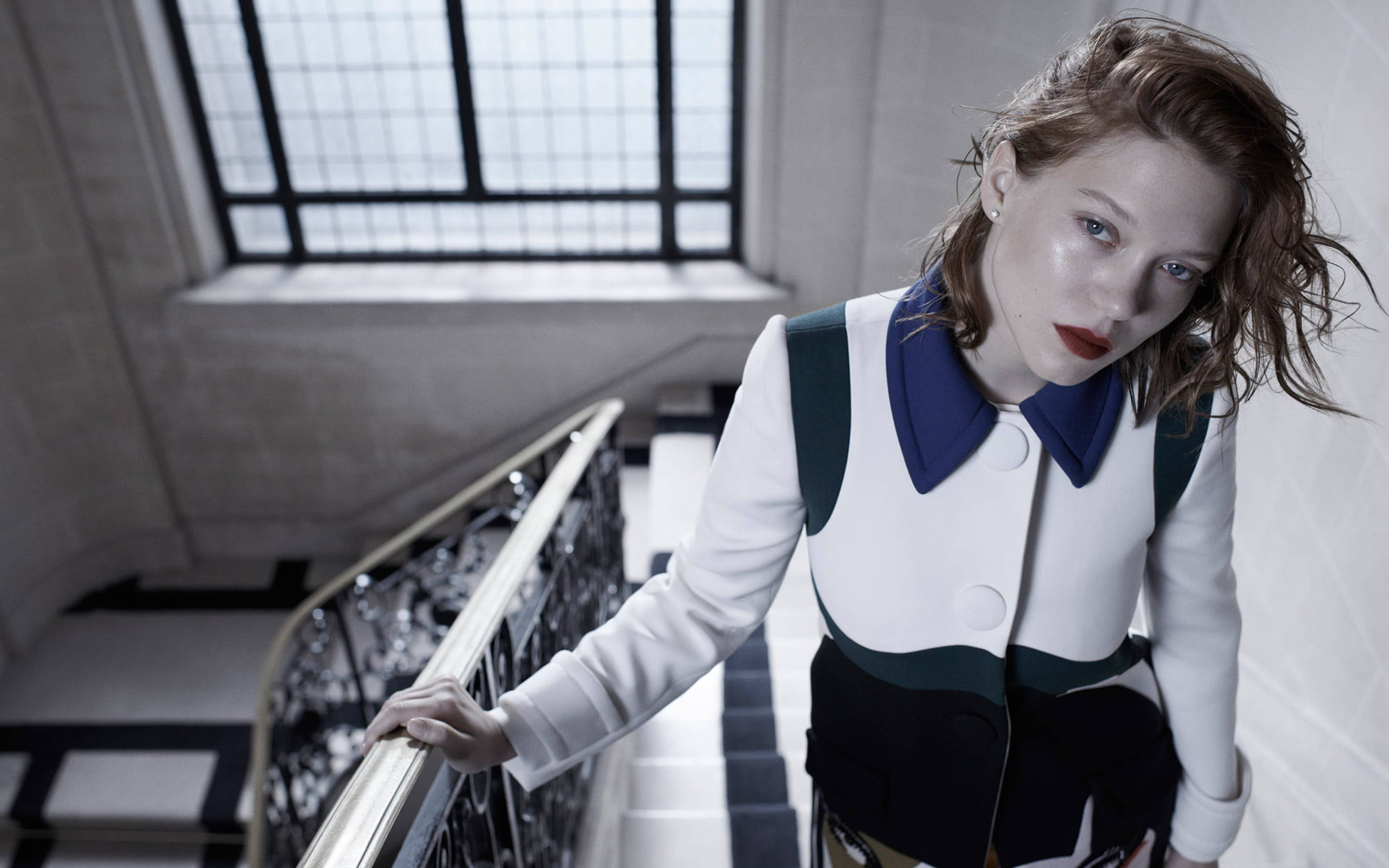 Lea Seydoux 桌布 壁紙 — 一位紅唇亂髮的女性站在宏偉的樓梯上，抓緊華麗的扶手，上方是帶格柵窗的天窗。
