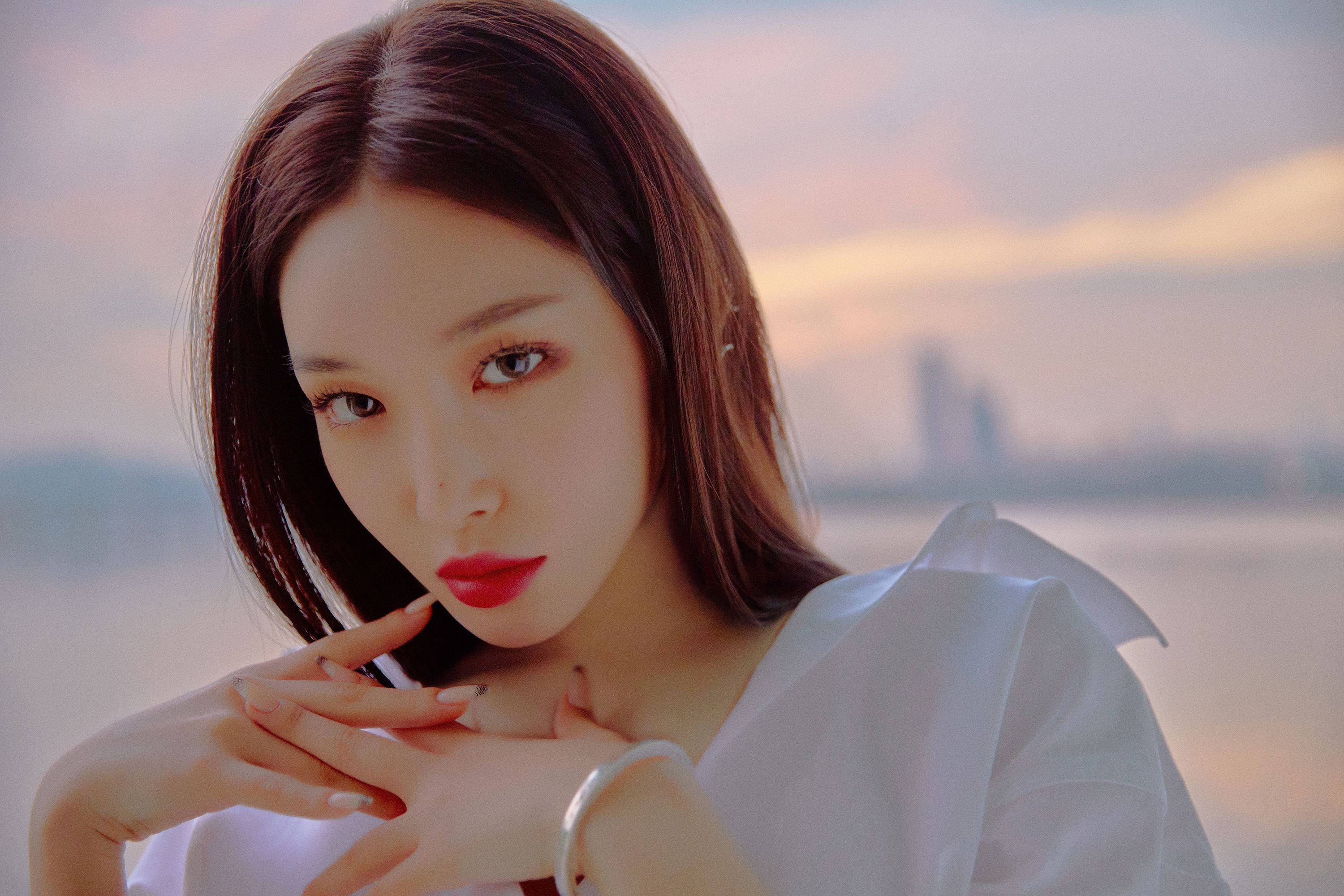 Chungha 桌布 壁紙 — 一位紅唇黑髮的女性溫柔地凝視著水上朦朧的日落，遠處有城市天際線。