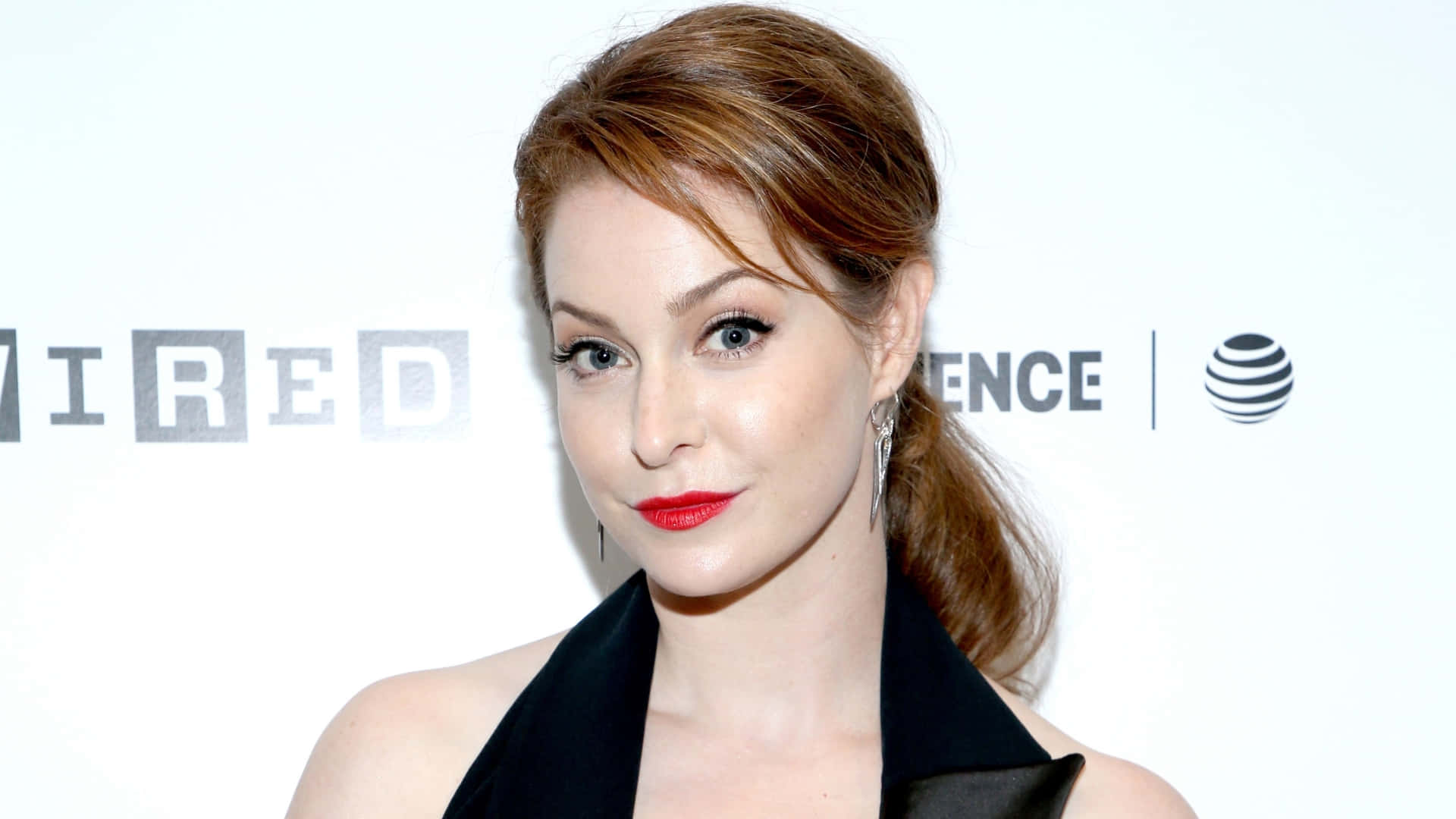 Esmé Bianco 桌布 壁紙 — 一位紅髮、紅唇大膽的女性，在白色活動背景前自信地穿著黑色連衣裙擺姿勢。