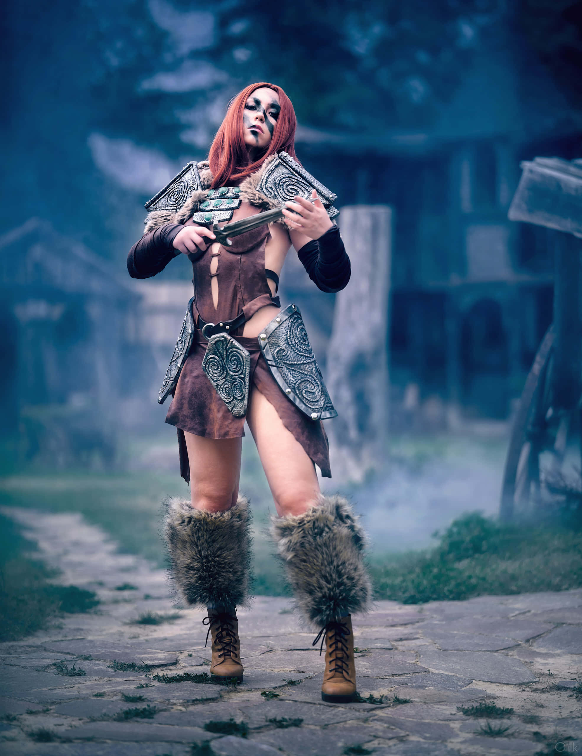Aela The Huntress 桌布 壁紙 — 一位紅髮、身穿華麗盔甲的女戰士，佇立在廢棄村莊霧氣瀰漫的石徑上。