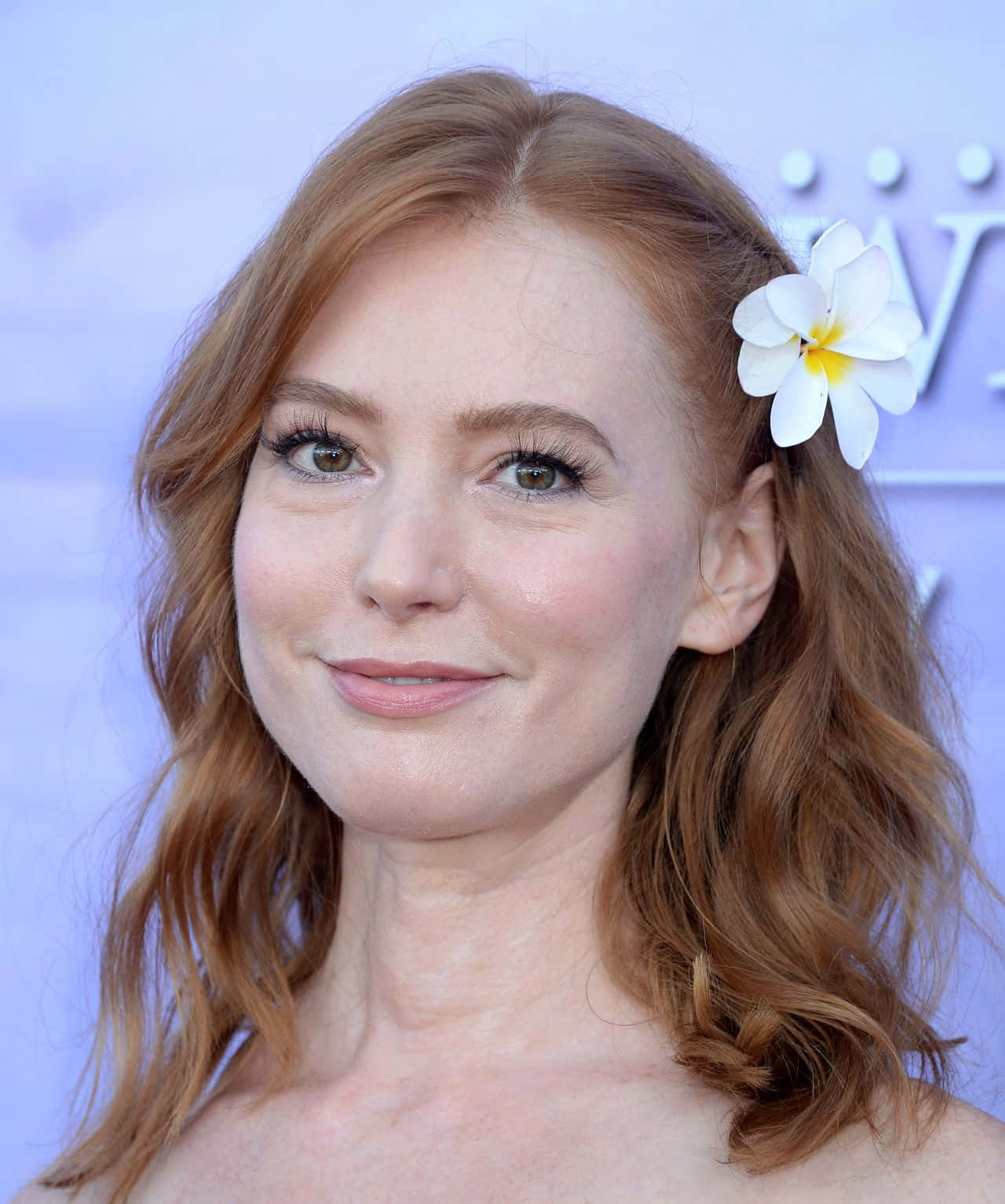 Alicia Witt 桌布 壁紙 — 一位紅髮女子，頭上戴著一朵白色雞蛋花，在淺藍色背景下溫柔微笑。