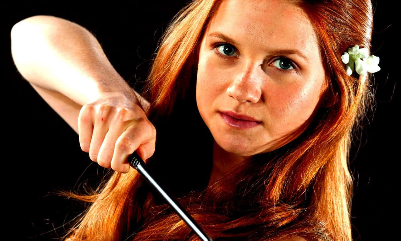 Ginny Weasley 桌布 壁紙 — 一位紅髮女子，頭髮上插著一朵白花，在黑色背景下緊握魔杖，眼神專注。
