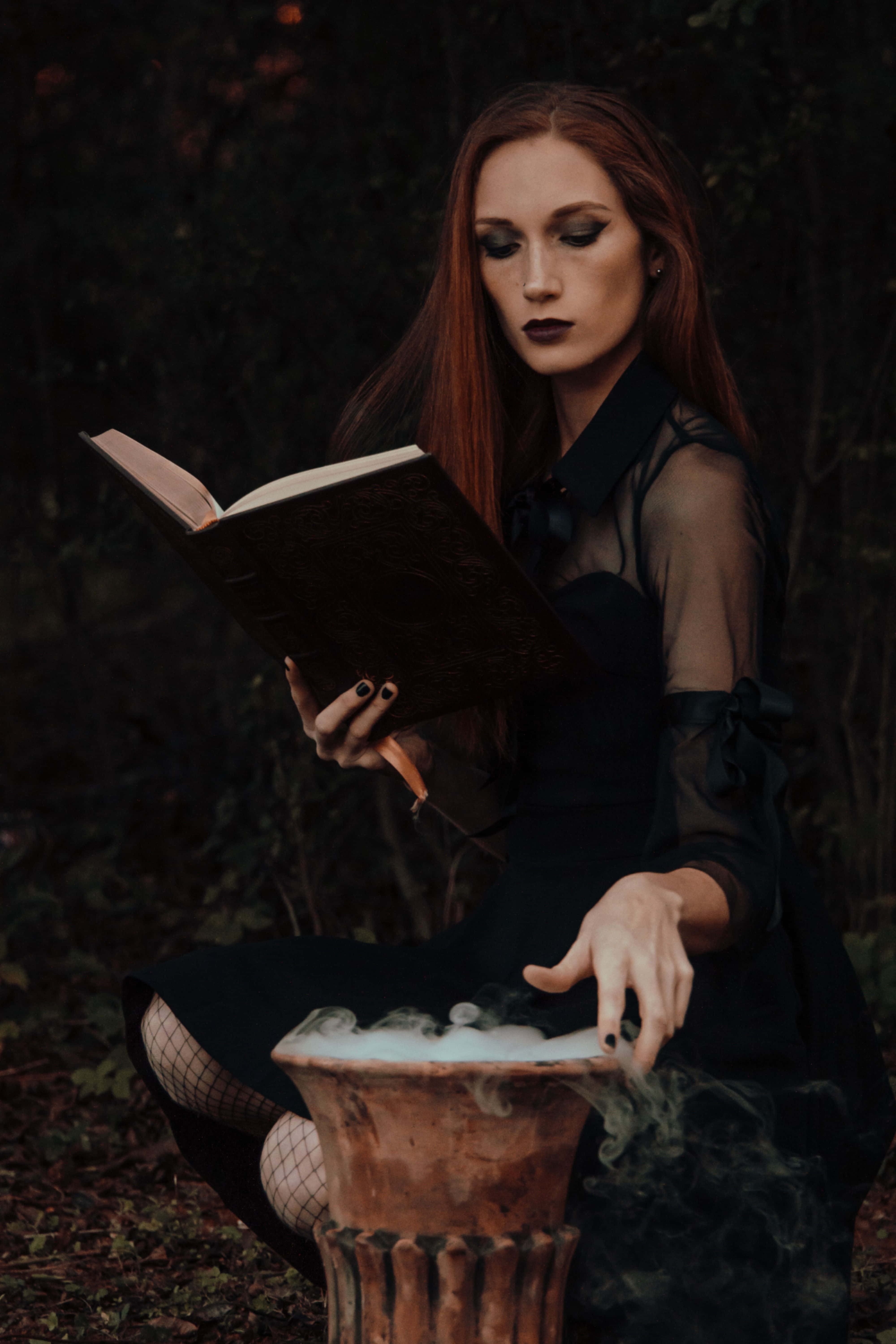 Witch Costumes 桌布 壁紙 — 一位紅髮女子在黑暗的森林中閱讀一本書，她的手懸在一個冒著蒸氣的大鍋上方。