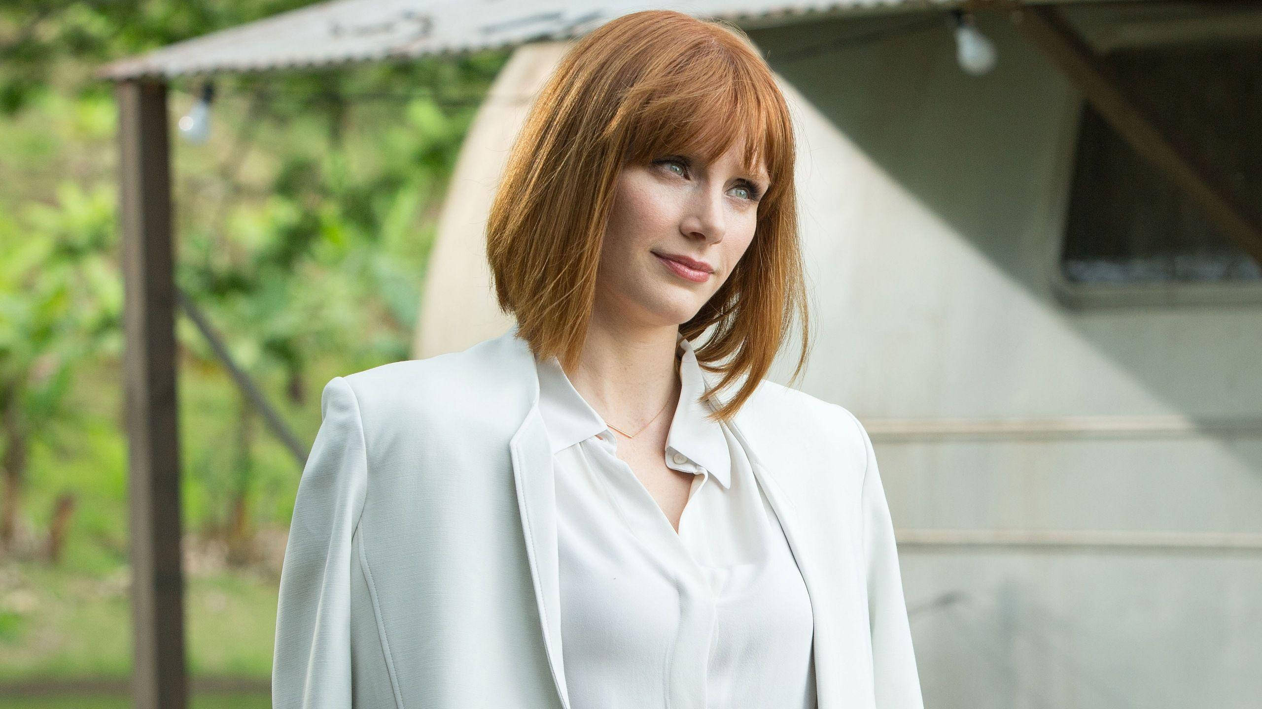 Jurassic World 桌布 壁紙 — 一位紅髮女子身穿白色西裝外套，站在戶外一輛拖車附近，身後有綠色植物。