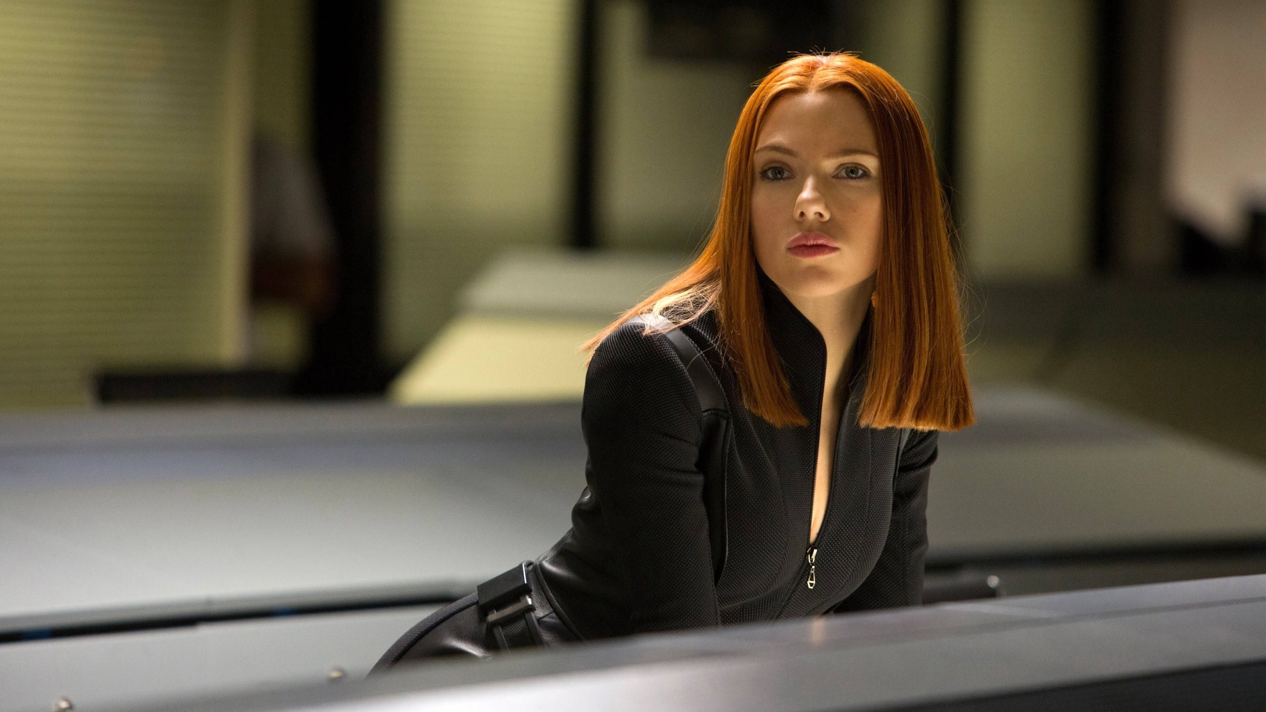 Scarlett Johansson Black Widow 桌布 壁紙 — 一位紅髮女子身穿黑色皮夾克，在燈光昏暗的辦公室裡俯身靠在一張桌子上。