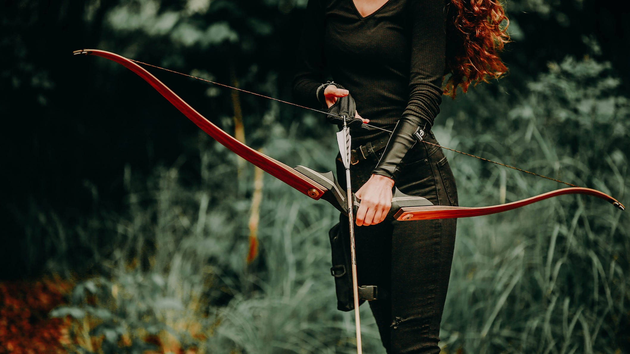 Archery 桌布 壁紙 — 一位紅髮女性在黑暗的森林中拉開紅色弓，周圍有高草和柔和的綠色色調。