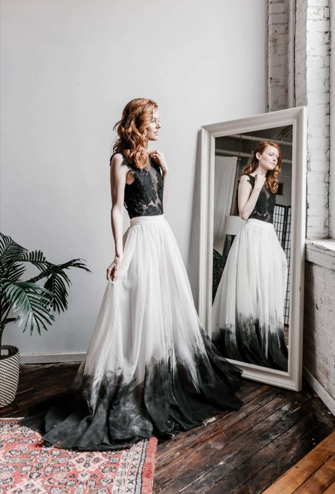 Black And White Wedding 桌布 壁紙 — 一位紅髮女性站在簡約風格的房間裡，身穿黑色蕾絲上衣和由白漸變至黑的裙子，凝視著窗邊高鏡中的倒影。