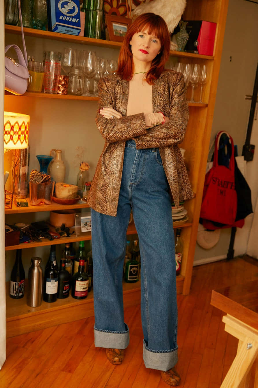 Woman 桌布 壁紙 — 一位紅髮女性自信地站在燈光溫暖的房間裡，穿著蛇皮西裝外套和寬腿牛仔褲，周圍是擺滿 eclectic 裝飾的架子。