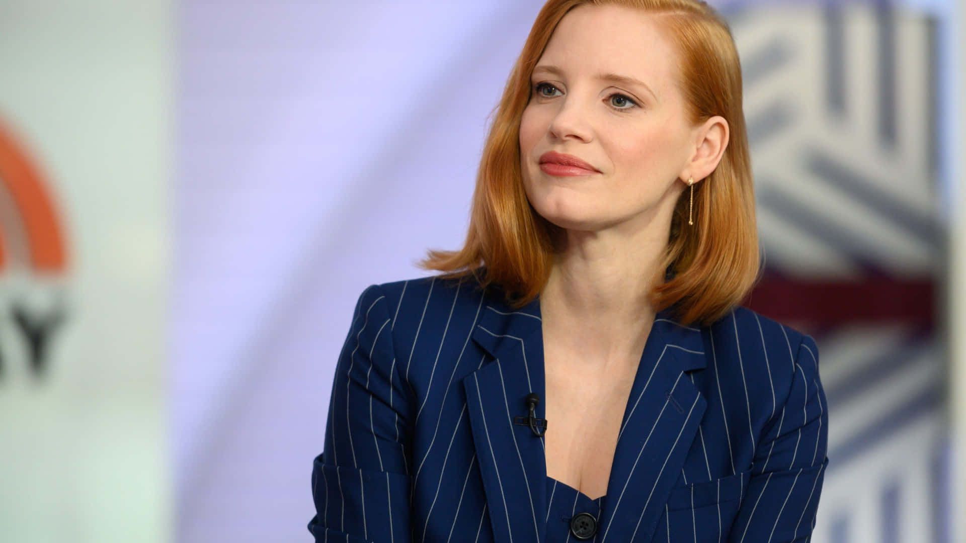 jessica chastain 桌布 壁紙 — 一位紅髮女性身穿藍色細條紋西裝外套，在模糊的攝影棚背景下若有所思地望向一旁。