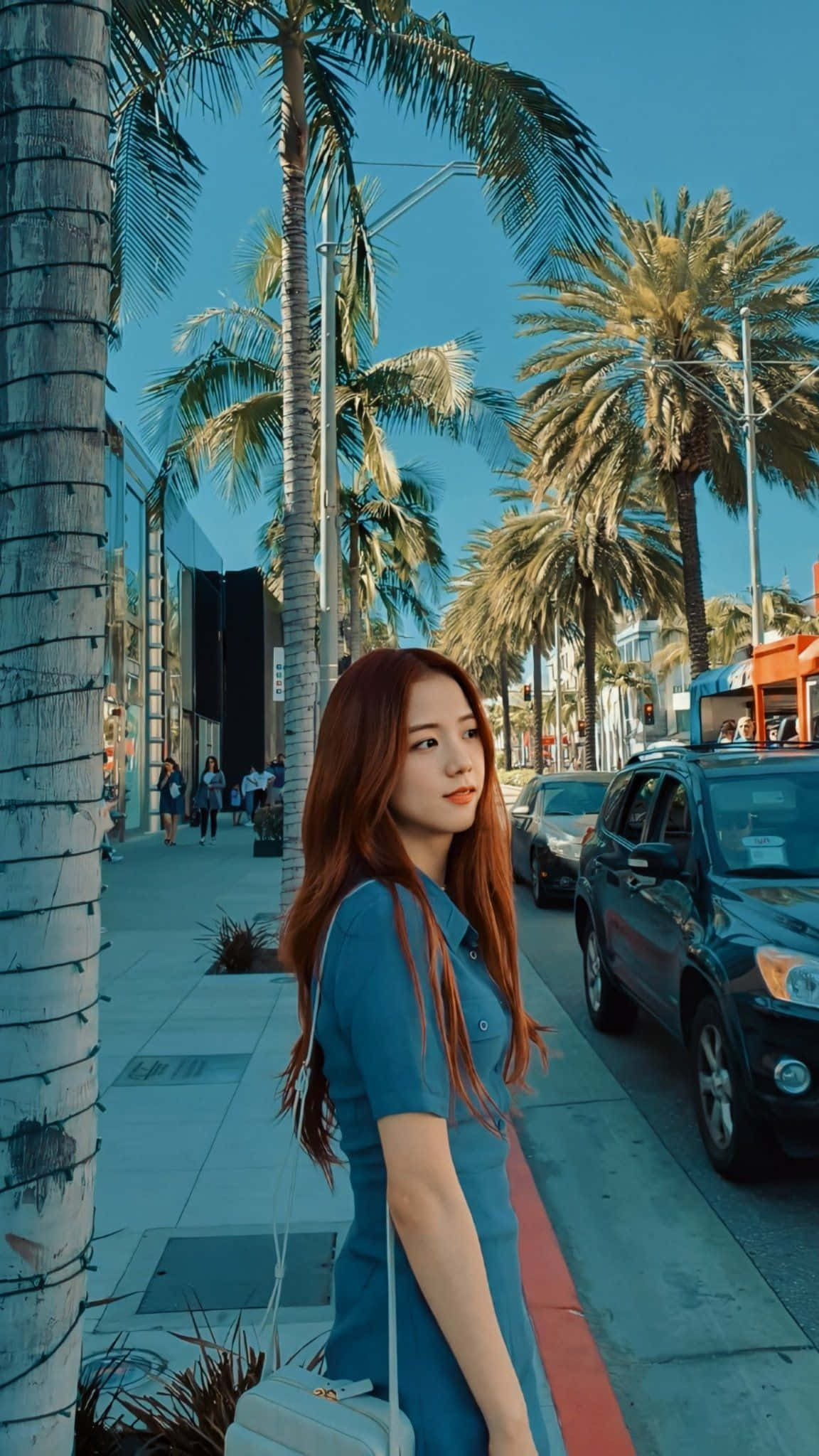 BLACKPINK 桌布 壁紙 — 一位紅髮年輕女子穿著藍色連衣裙，站在棕櫚樹成行的人行道上，頭頂是明亮的藍天。