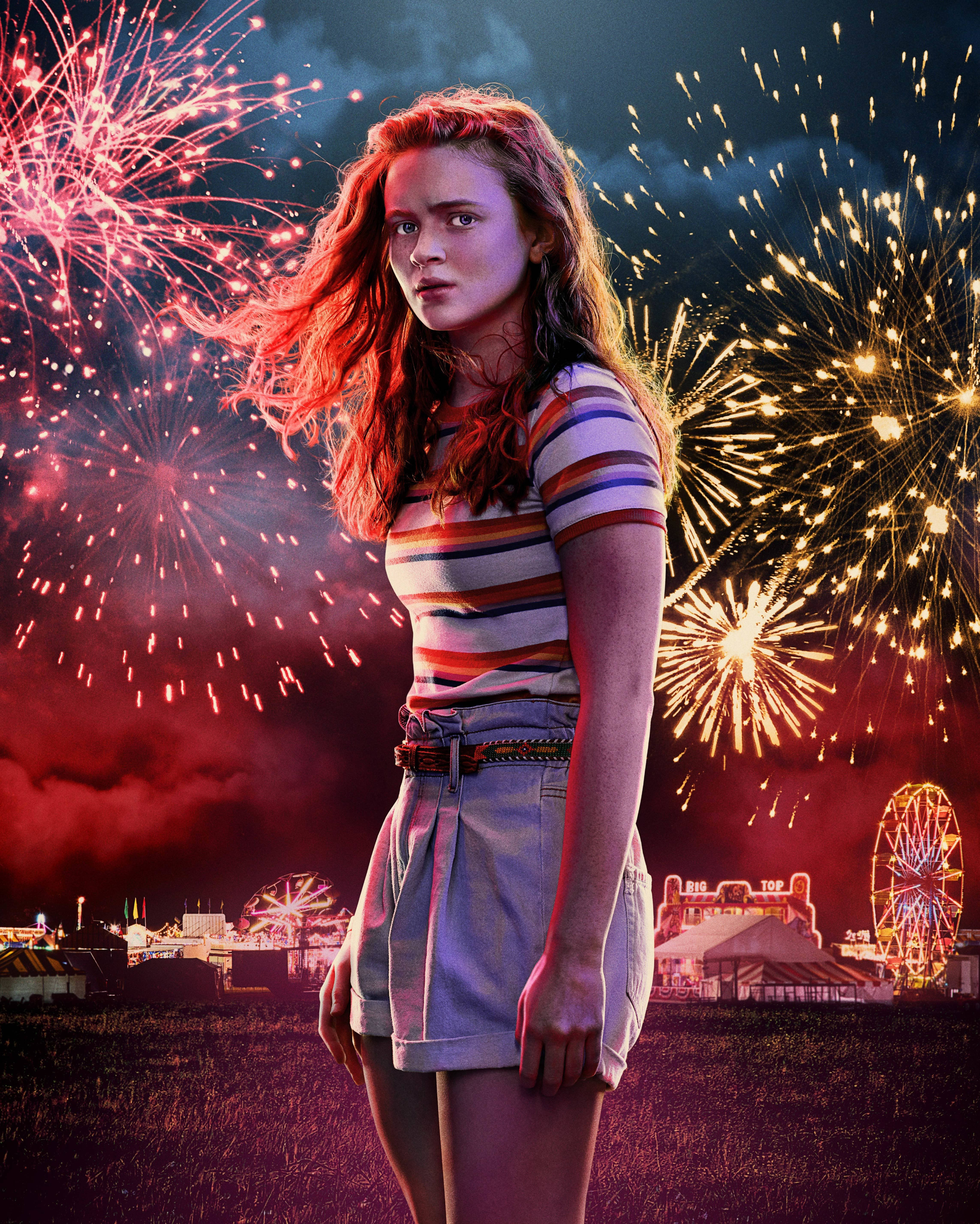 Stranger Things 3 桌布 壁紙 — 一位紅髮年輕女子站在田野中，夜晚嘉年華上空綻放著五彩繽紛的煙火。