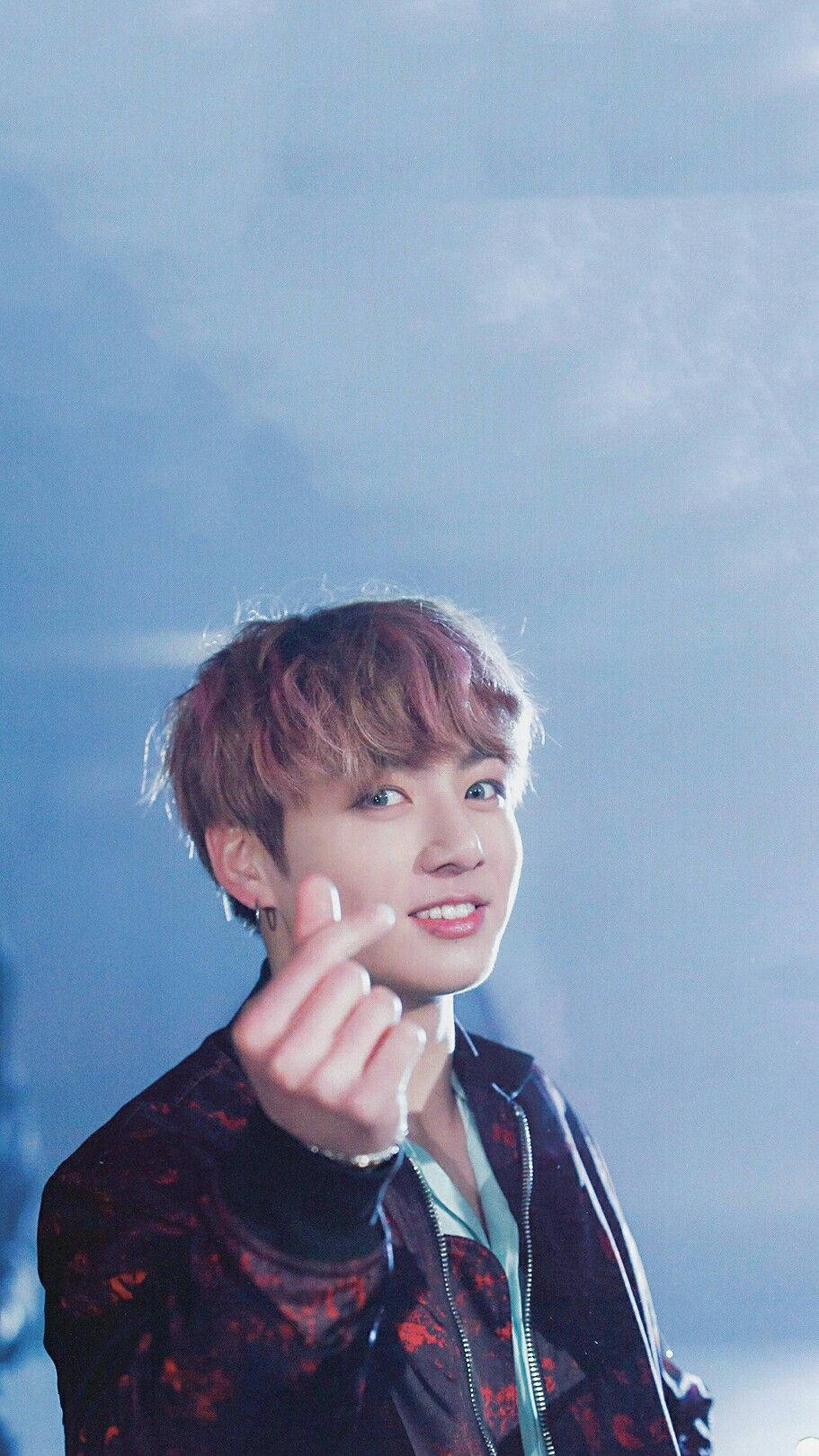 Jungkook 桌布 壁紙 — 一位紅髮年輕男子在柔和的藍色模糊背景前微笑著做出手指愛心手勢。