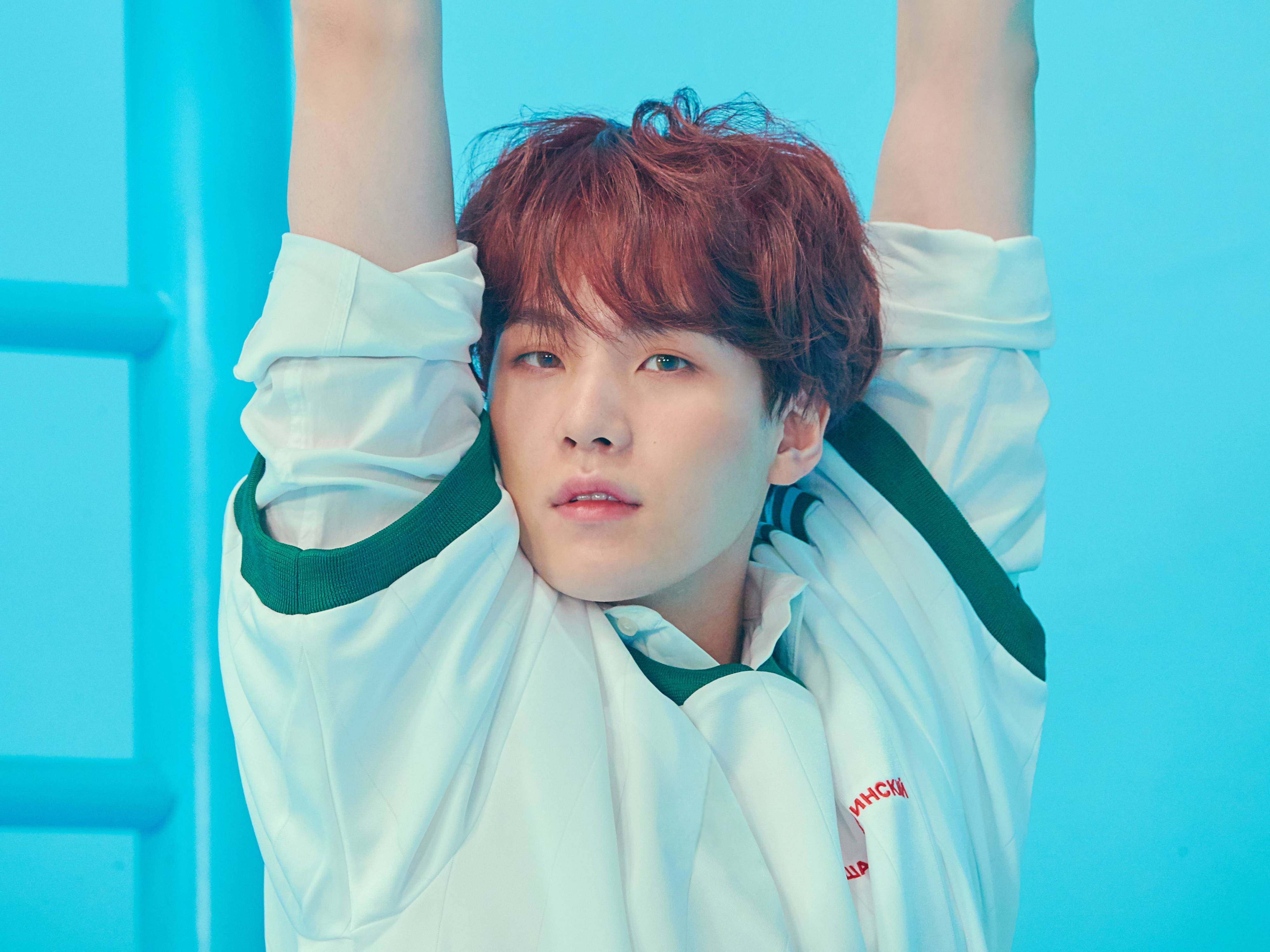 슈가 BTS 桌布 壁紙 — 一位紅髮年輕男子穿著白色外套，在鮮豔的藍色背景前向上伸展雙臂。