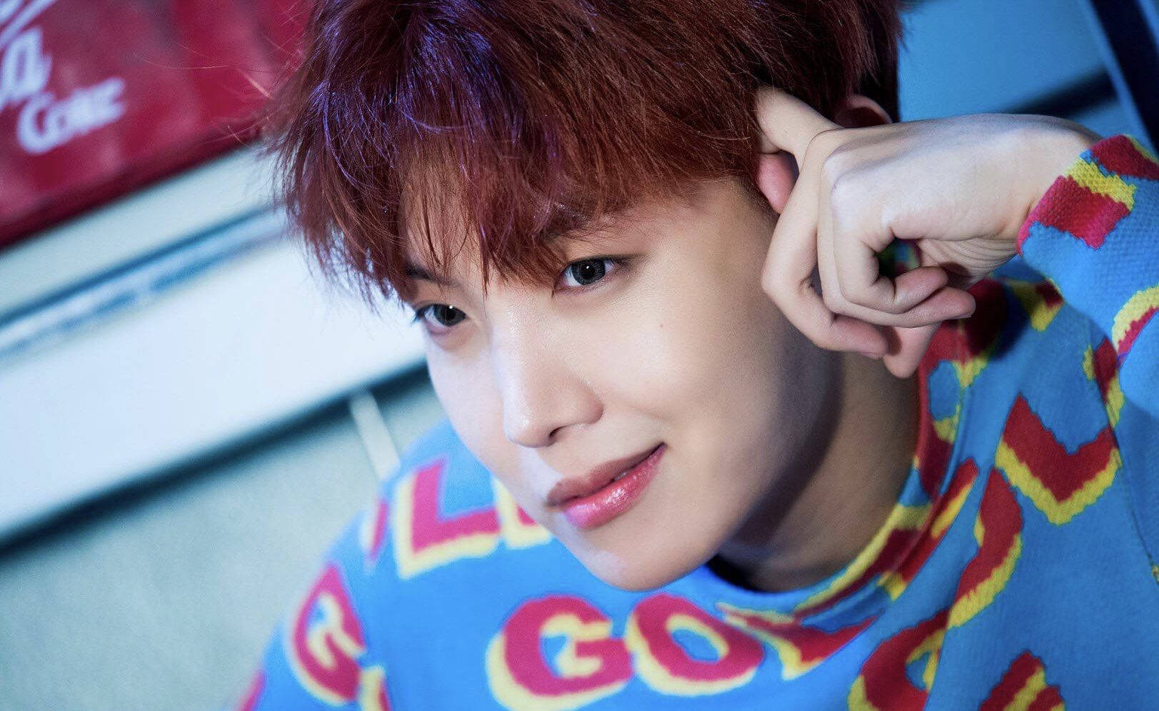 jhope 桌布 壁紙 — 一位紅髮年輕男子穿著色彩繽紛的毛衣，將手放在耳邊，溫柔地凝視著。