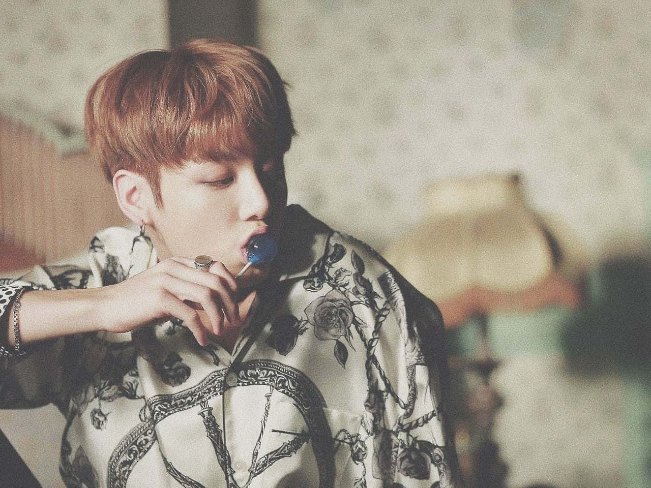 Jungkook 桌布 壁紙 — 一位紅髮年輕男子穿著花卉圖案襯衫，在柔和光線的房間裡舔著藍色棒棒糖。