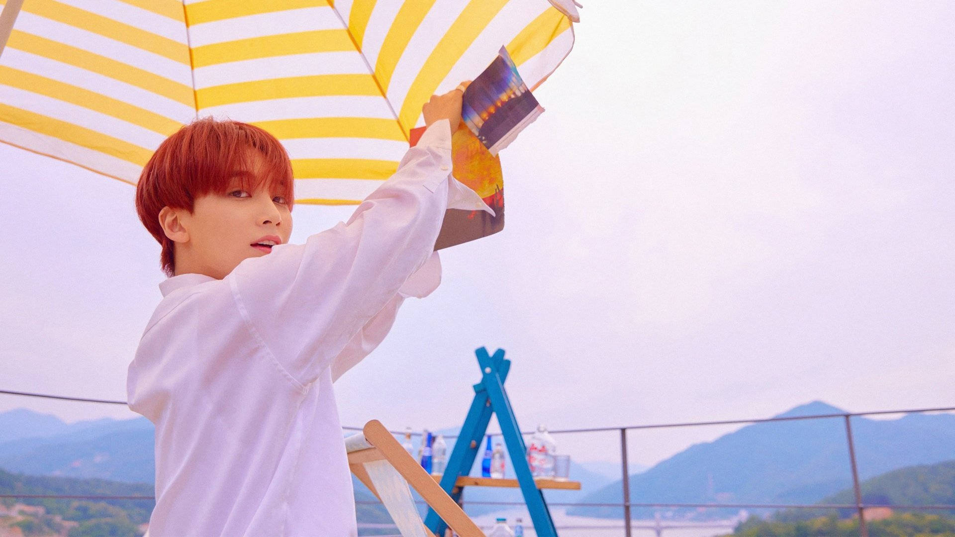 yoon jeonghan 桌布 壁紙 — 一位紅髮年輕男子站在陽台上，俯瞰著柔和天空下的山脈，手持一把黃白條紋雨傘。