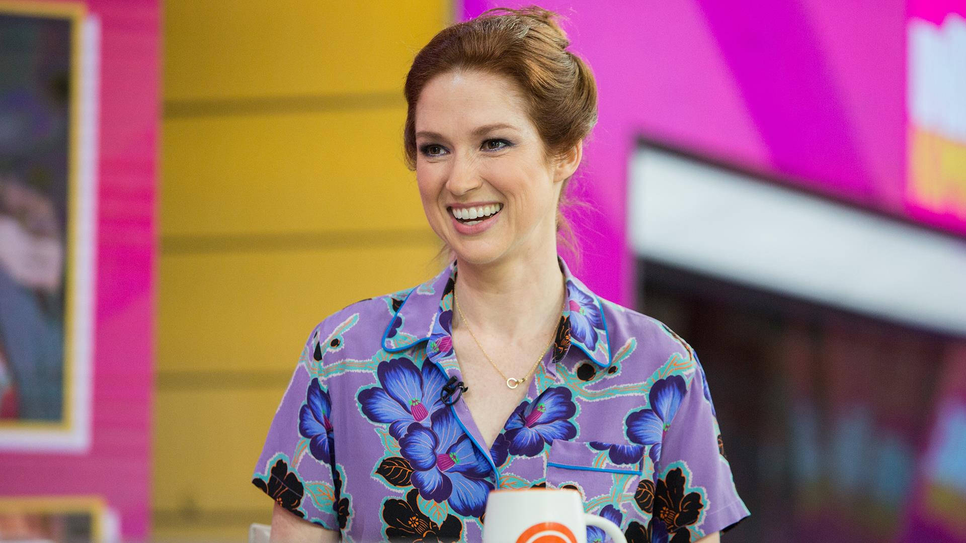 unbreakable kimmy schmidt 桌布 壁紙 — 一位紅髮微笑的女子身穿紫色花卉襯衫，在充滿活力的粉紅和黃色工作室背景前拿著一個馬克杯。