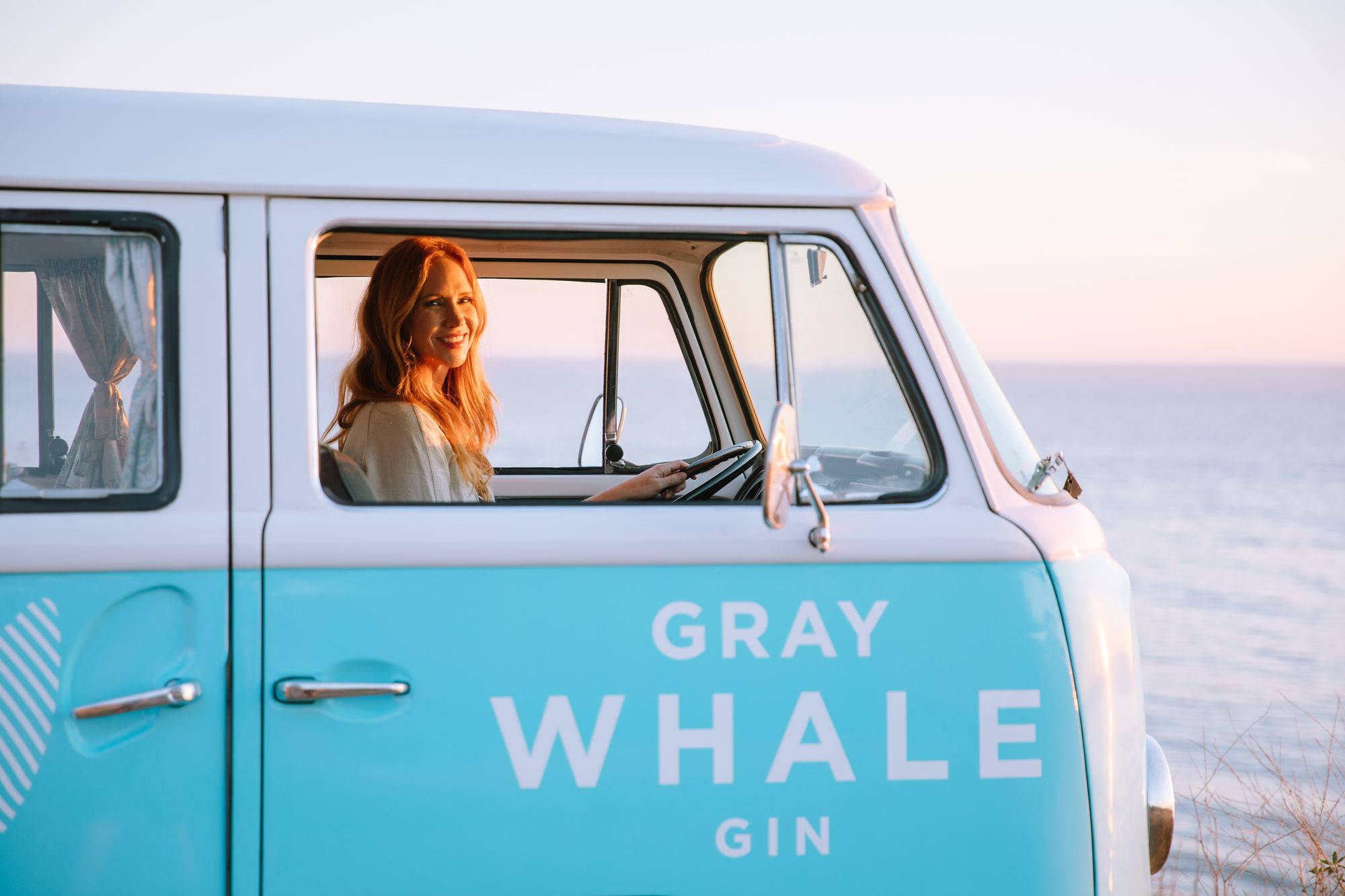 alcohol 桌布 壁紙 — 一位紅髮微笑的女子駕駛著一輛標有gray whale gin的藍綠色和白色復古廂型車，沿著日落時分的海岸道路行駛。