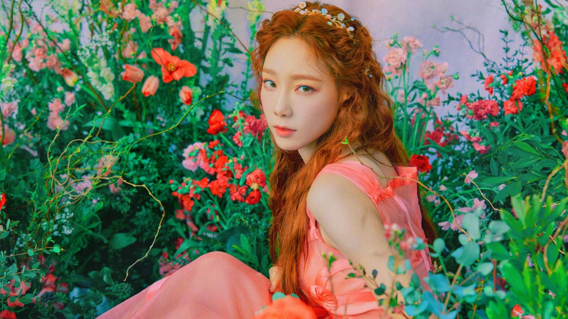 Taeyeon 桌布 壁紙 — 一位紅髮戴花冠的女性坐在茂密的綠植和鮮豔的紅粉色花朵中，身穿飄逸的桃色連衣裙。