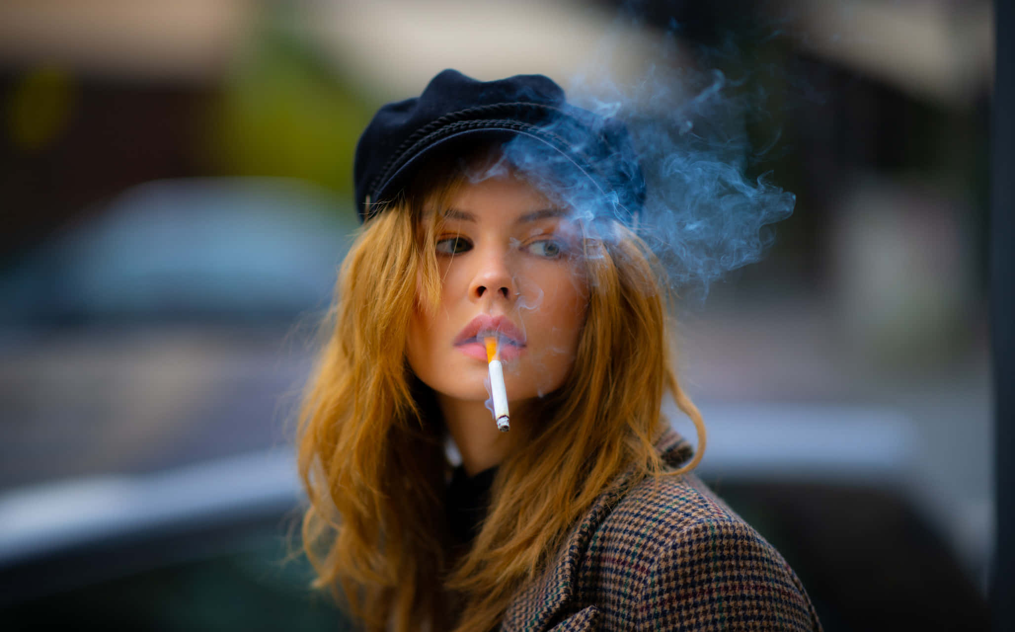 Girl Smoking 桌布 壁紙 — 一位紅髮戴黑色鴨舌帽的女性正在抽煙，煙霧從她的嘴唇繚繞升起，背景是模糊的城市景觀。