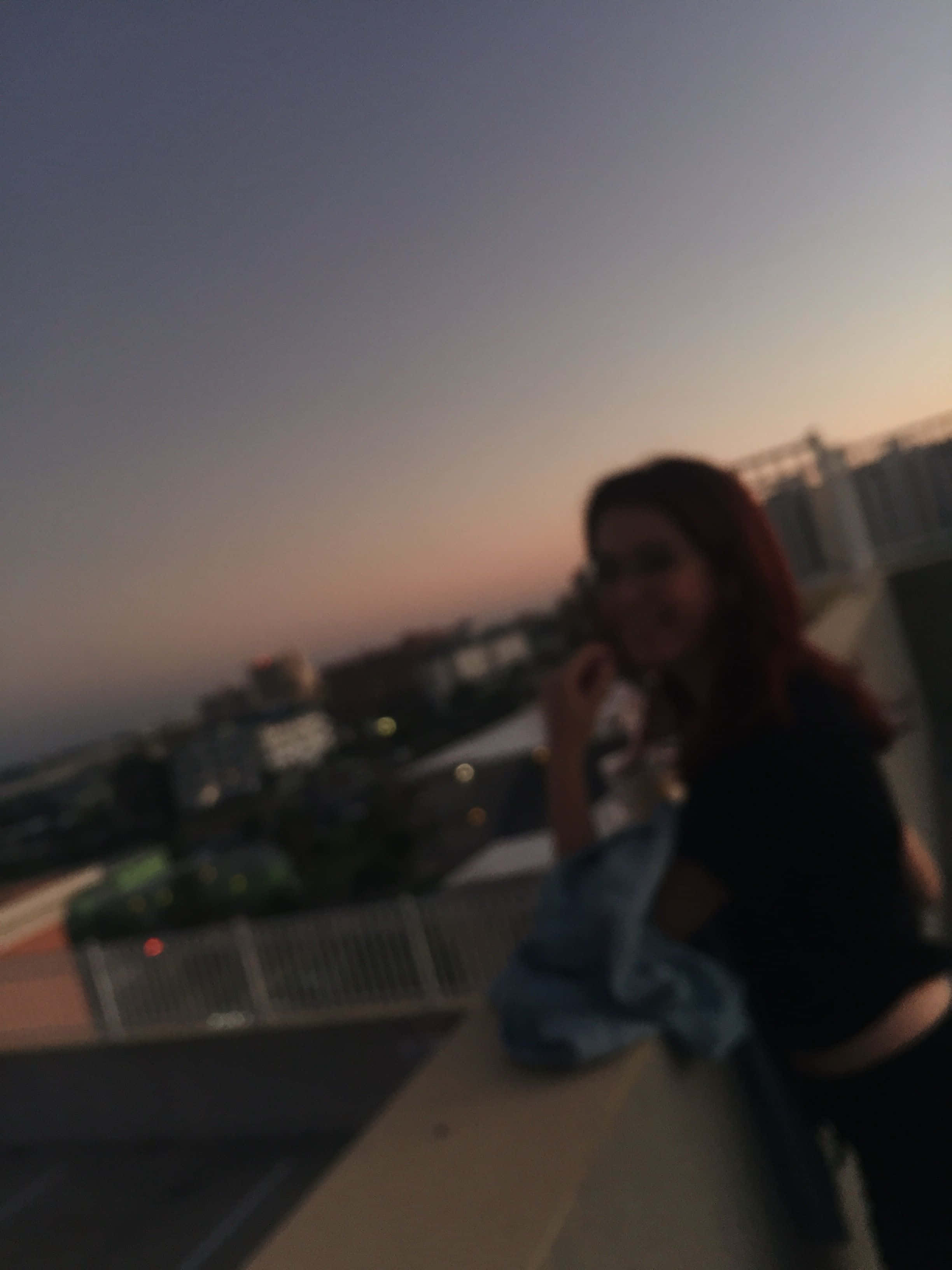 美學個人檔案 桌布 壁紙 — 一位紅髮的模糊女性在黃昏時分於屋頂上微笑，背後是城市景觀。