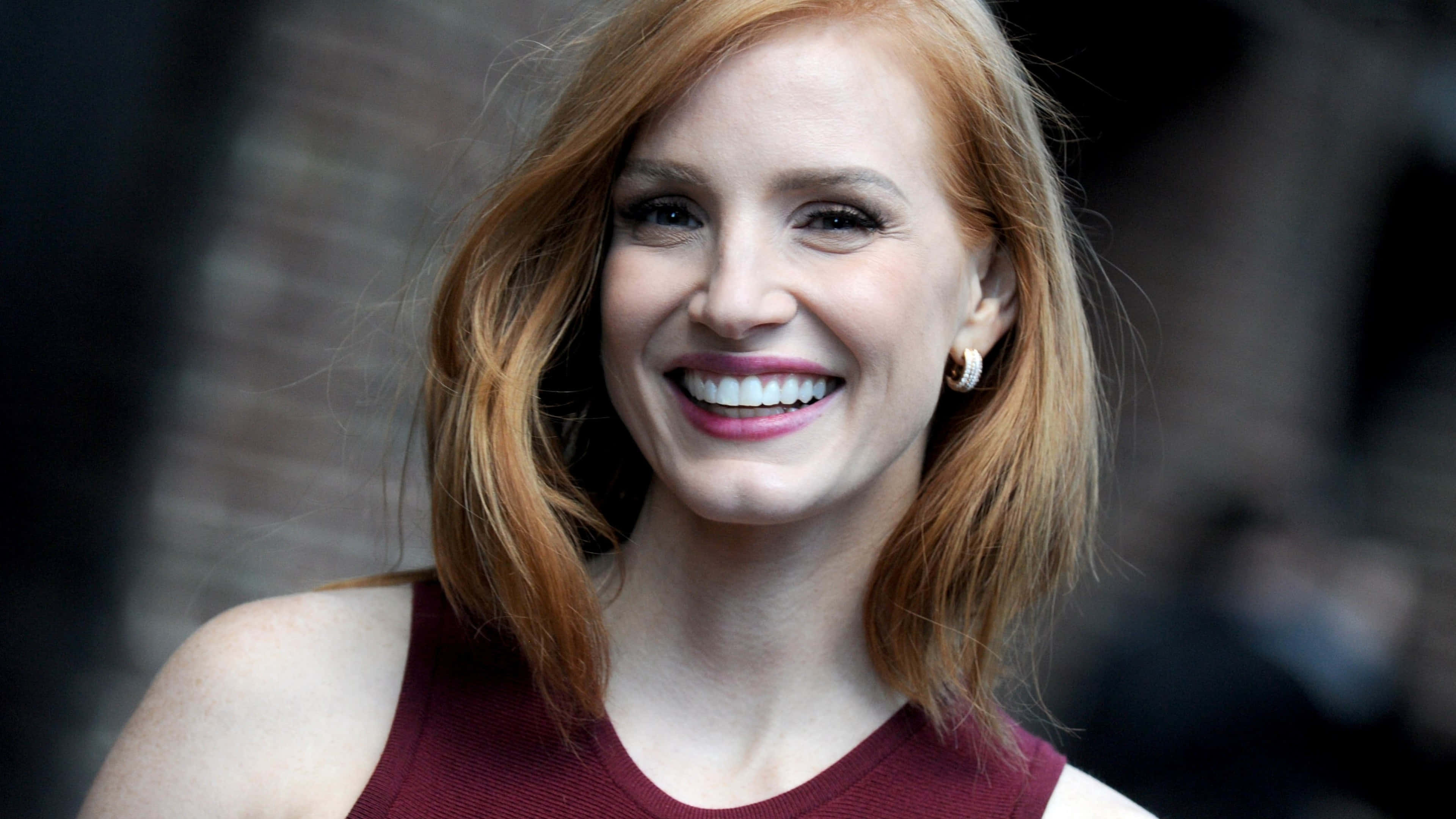 jessica chastain 桌布 壁紙 — 一位紅髮的燦爛女性，穿著栗紅色上衣，在模糊的磚牆背景下燦爛微笑。
