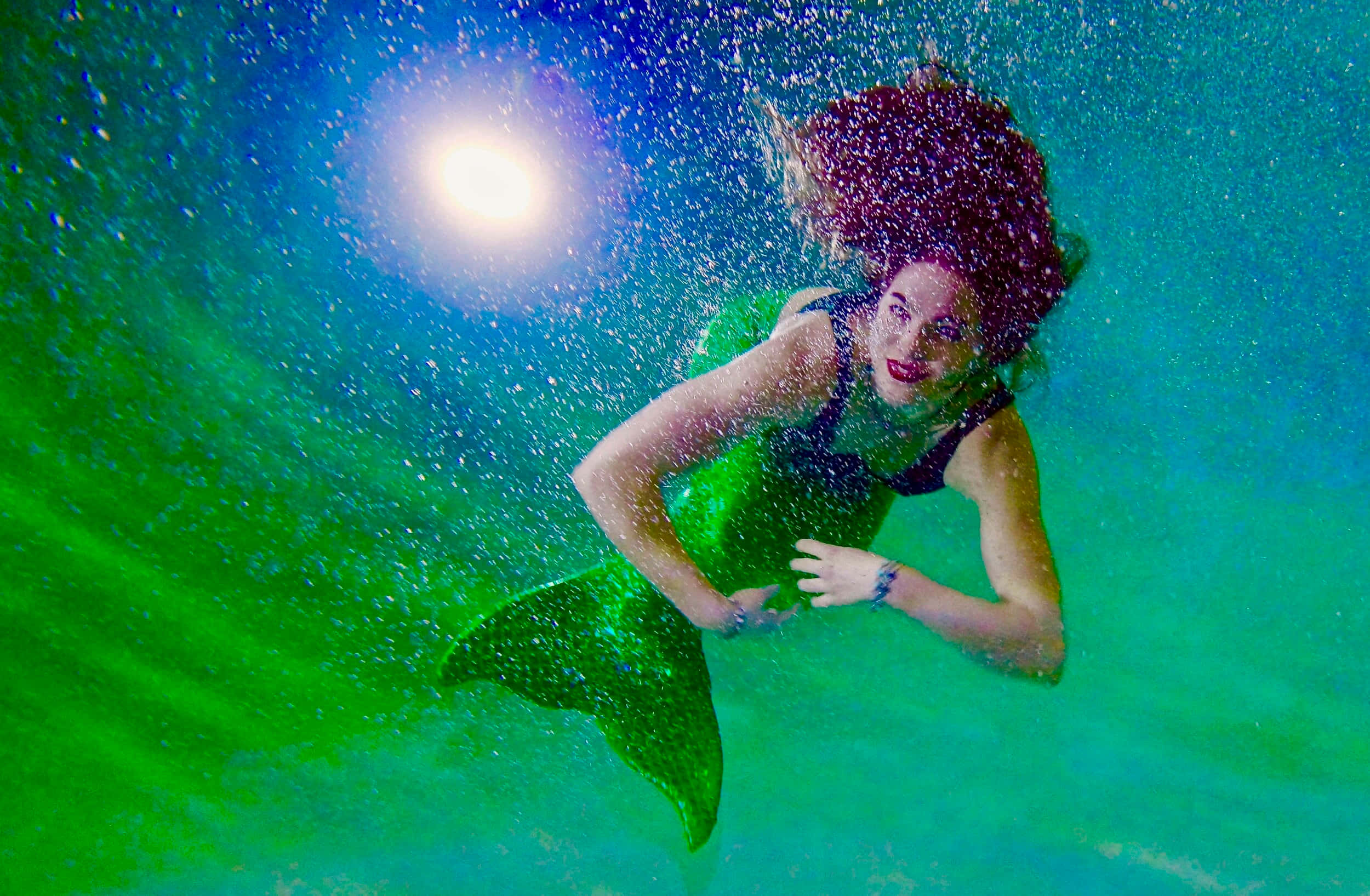 Mermaids 桌布 壁紙 — 一位紅髮綠尾的美人魚女子在碧綠的海水中游向鏡頭，陽光透過水面灑落。